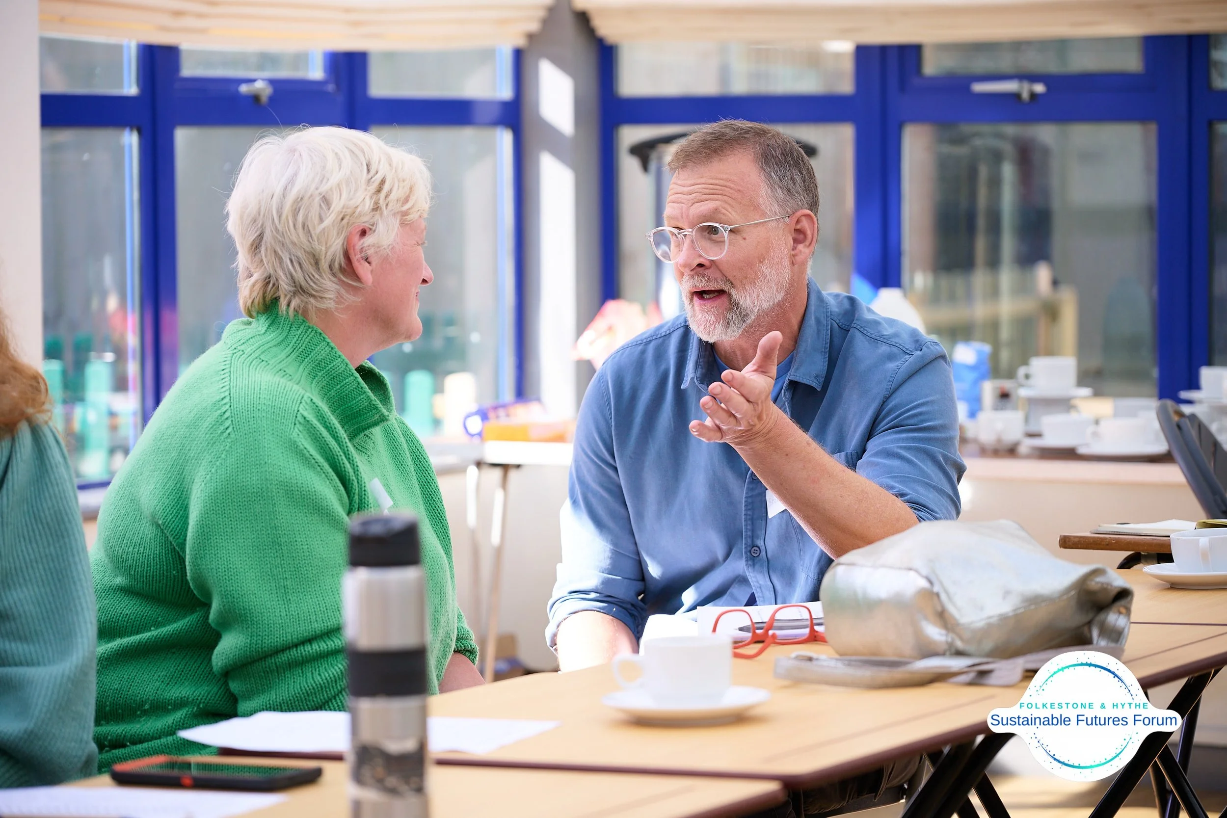 Two people having a conversation at a table in a bright room with large windows, one with blonde hair wearing a green sweater and the other with glasses and a beard wearing a blue shirt.