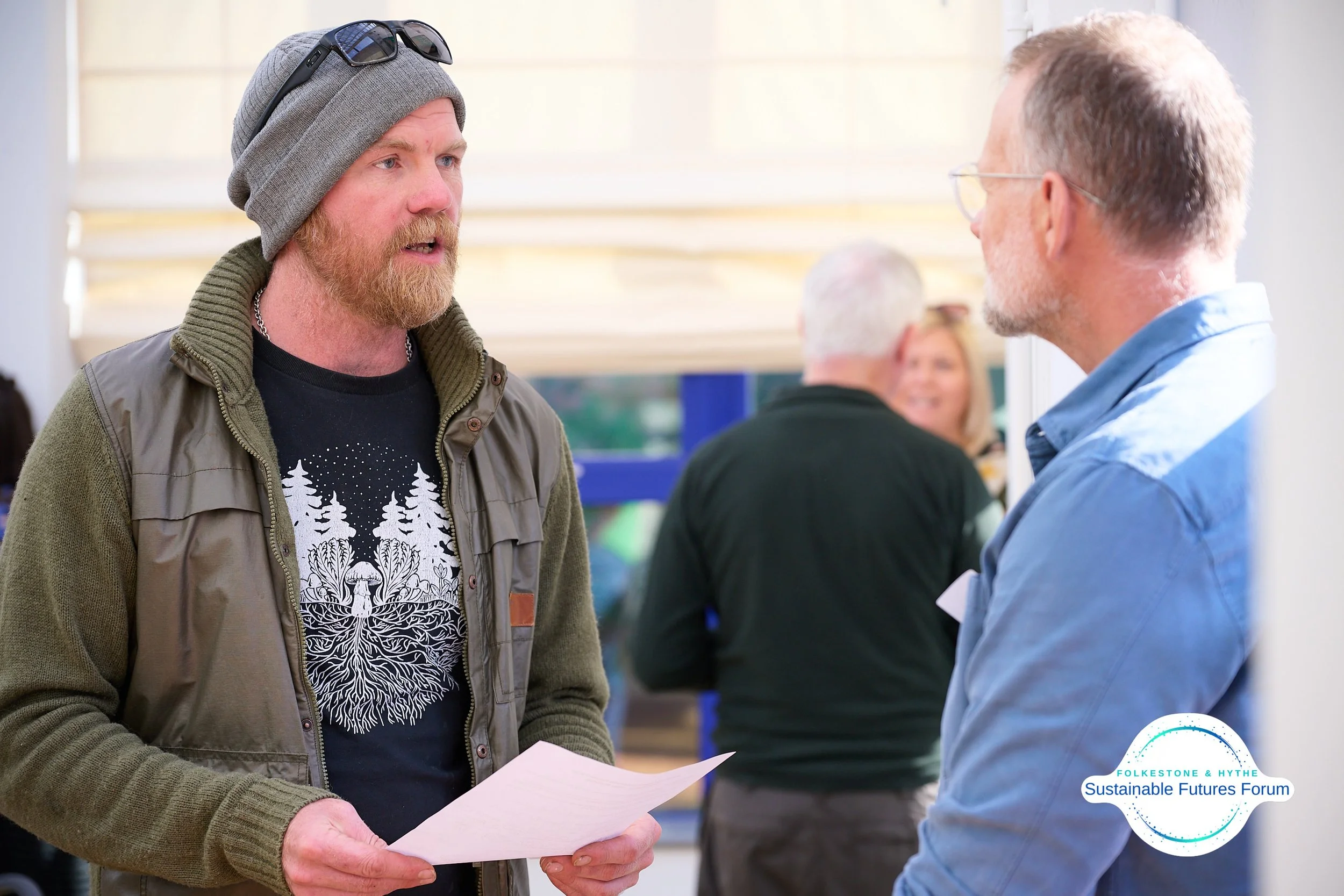 Two men are having a discussion at an indoor event, with people in the background. One man has a beard, is wearing a beanie, sunglasses, a jacket, and holding a paper. The other man has gray hair, glasses, and is wearing a blue shirt. There is a logo