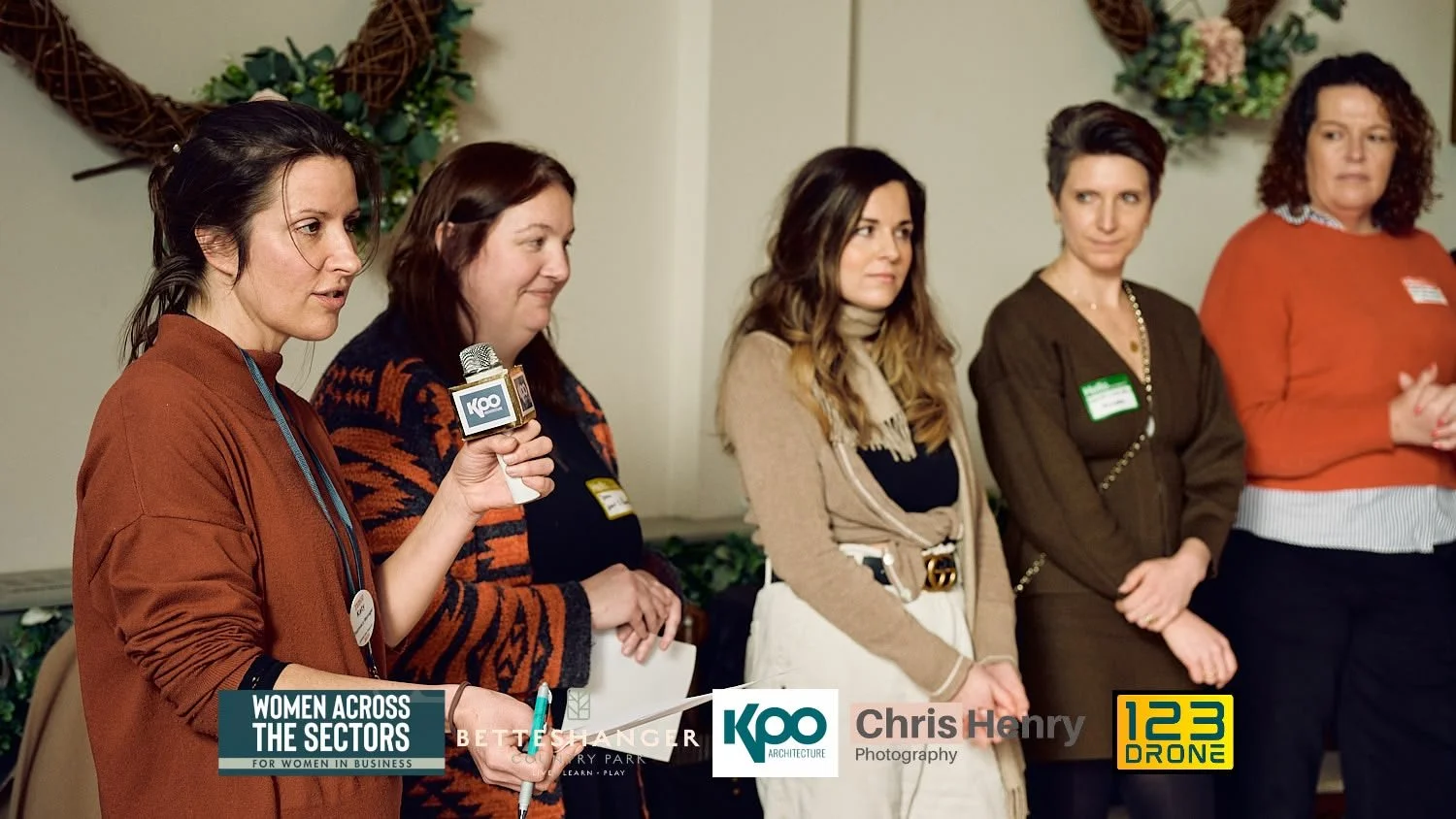 Five women standing in a row at a panel or event, one woman holding a microphone with the KAO logo, with a decorative wreath on the wall behind them.