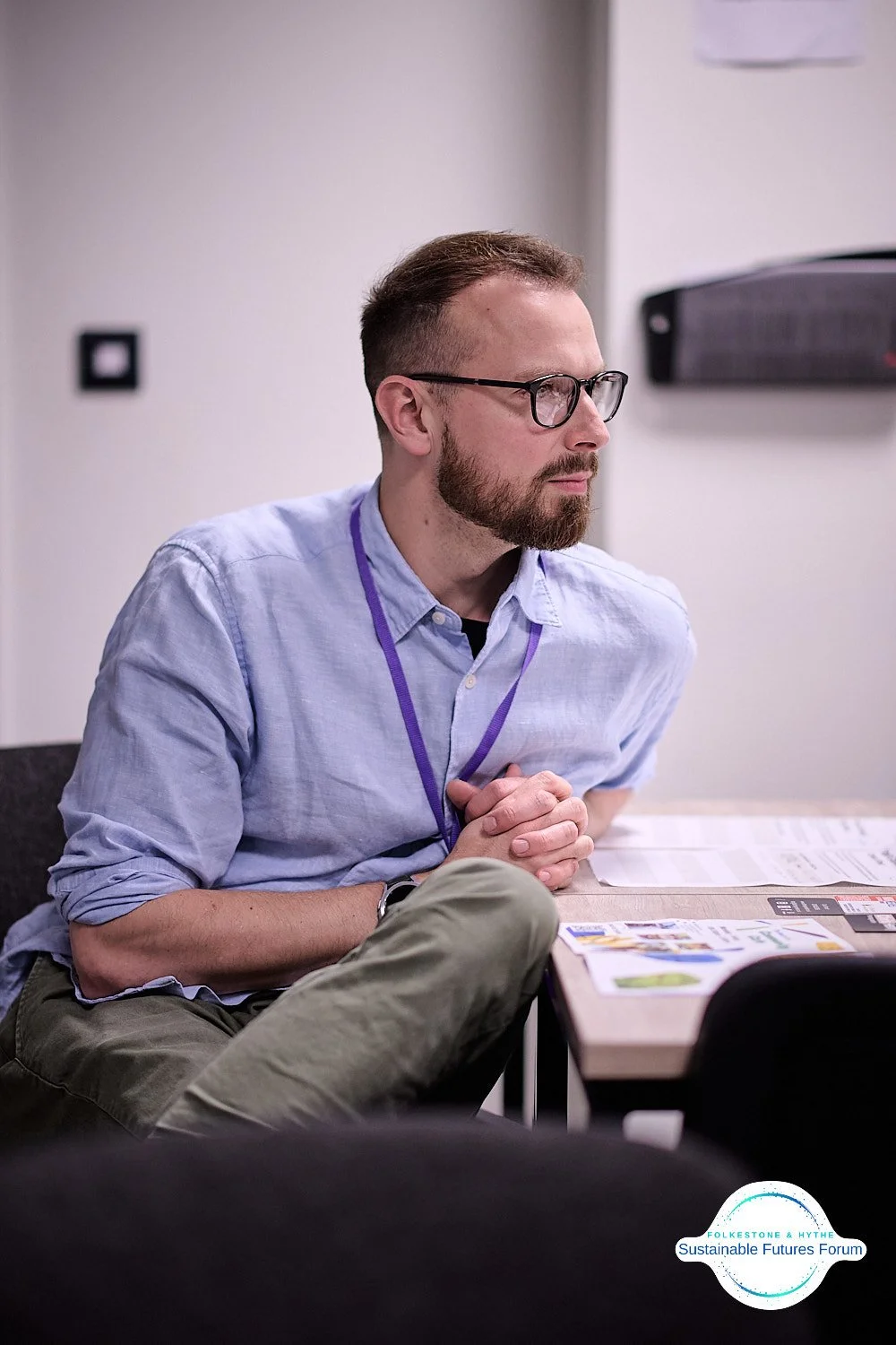 A man with glasses and a beard sits at a table during a meeting or workshop, with documents and a calculator on the table. A logo with the text 'Folketone & Aythe Sustainable Futures Forum' is visible in the bottom right corner.