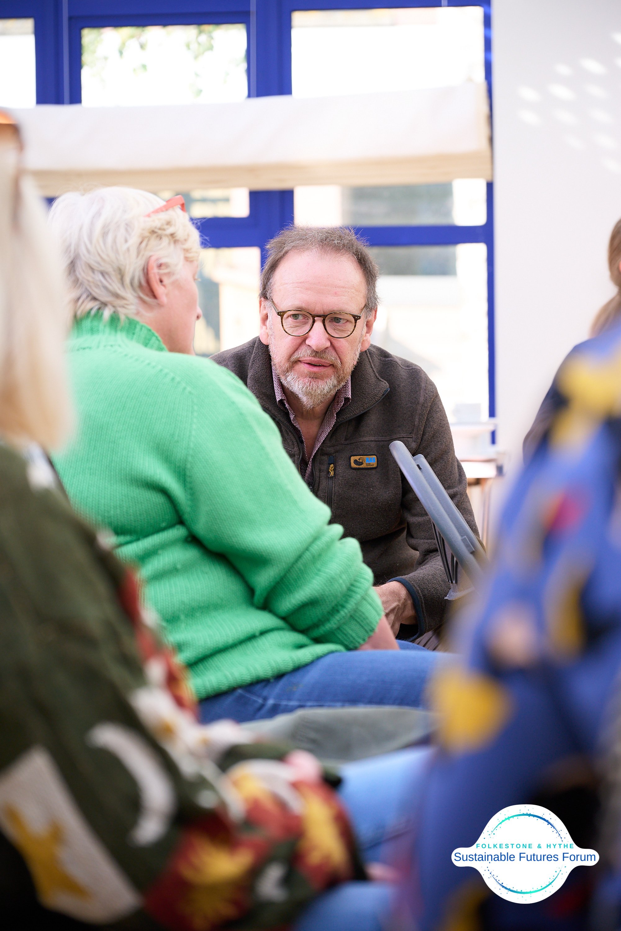 A man with glasses and a beard is talking to a woman with short gray hair and a bright green sweater, while others look on. They are seated in a room with bright natural light and blue-framed windows. There is a logo in the bottom right corner that s