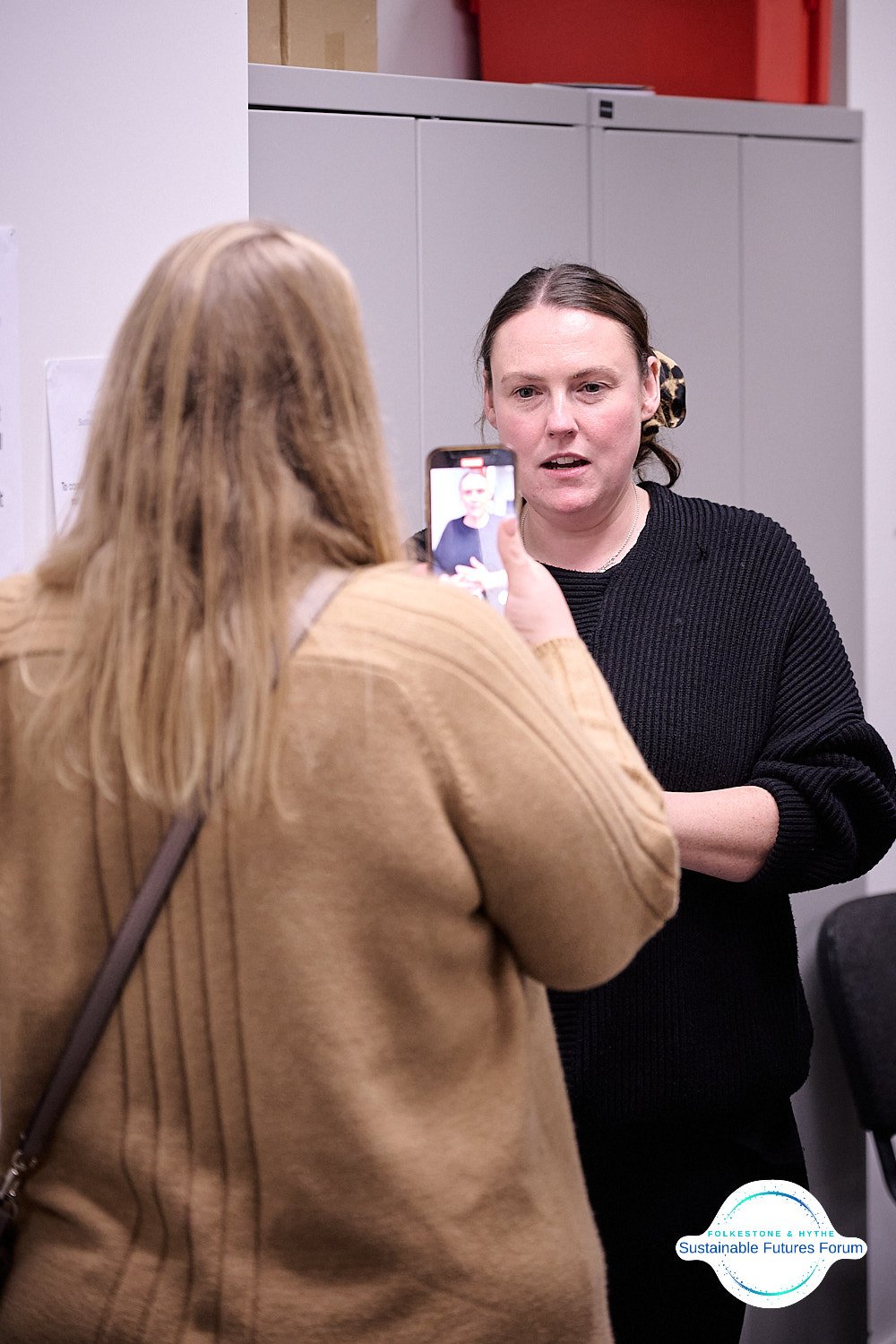 Two women are having a conversation, one is taking a photo of the other with her phone. The woman being photographed has dark hair in a ponytail with a leopard print scrunchie, wearing a black sweater. The photo appears to be taken indoors, with gray