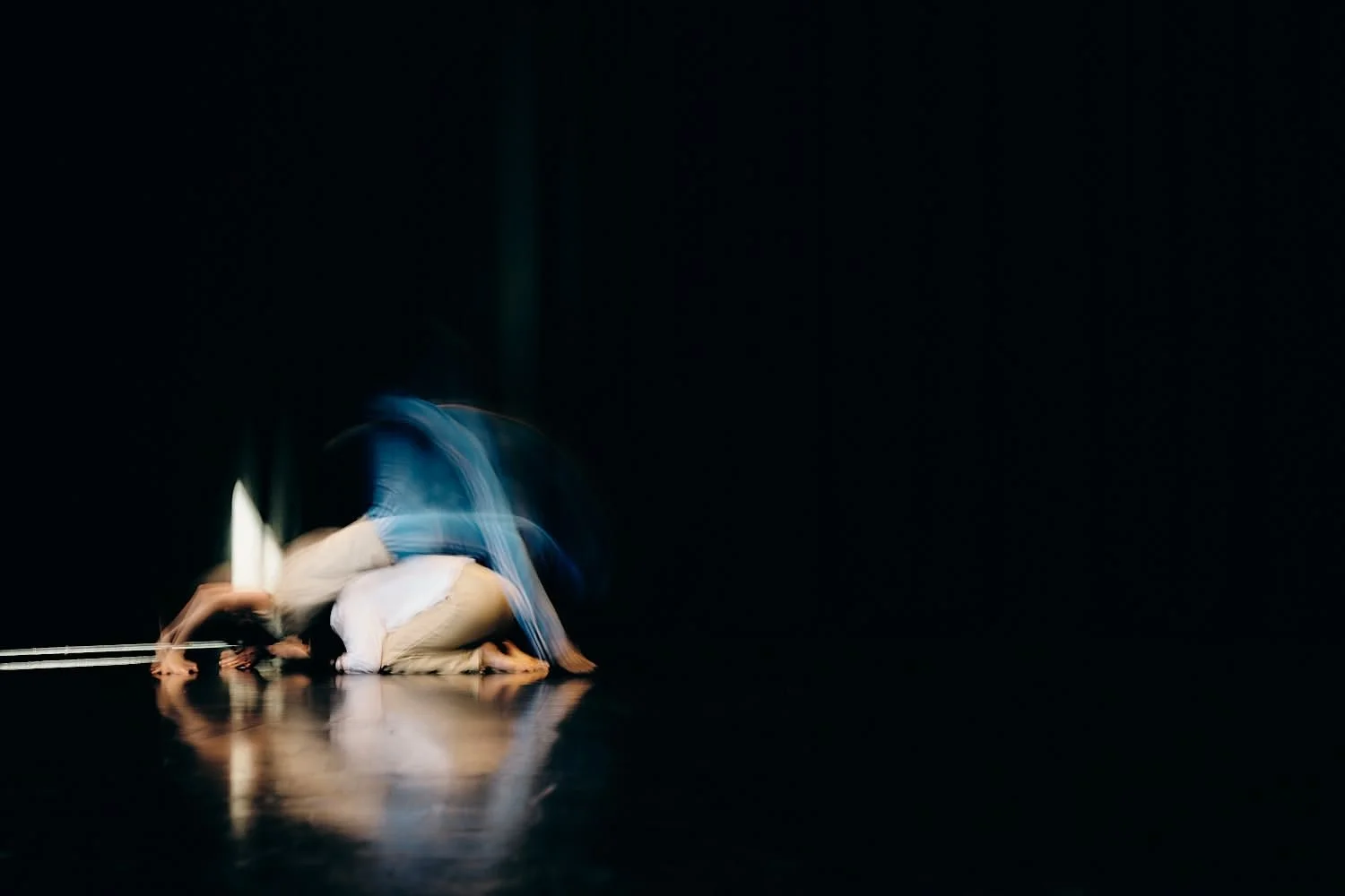 Two people are engaged in a dynamic dance or acrobatic performance on a dark stage with minimal lighting, creating a blur effect that emphasizes movement.