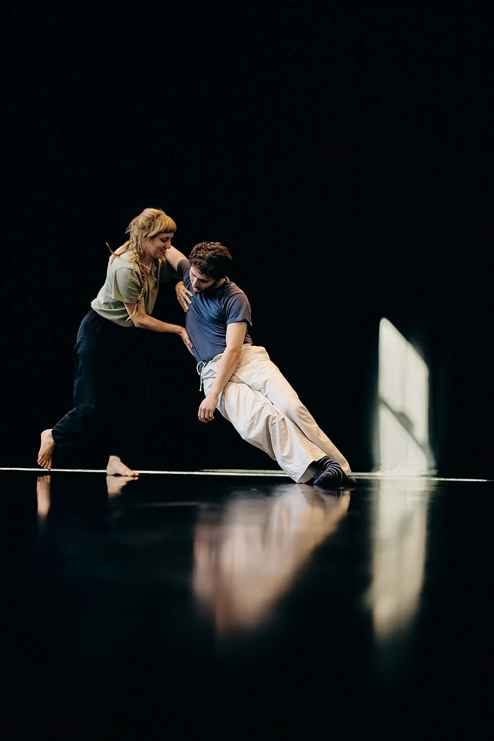 Two performers practicing dance or theater on a black stage with minimal lighting, one supporting the other who is leaning sideways.
