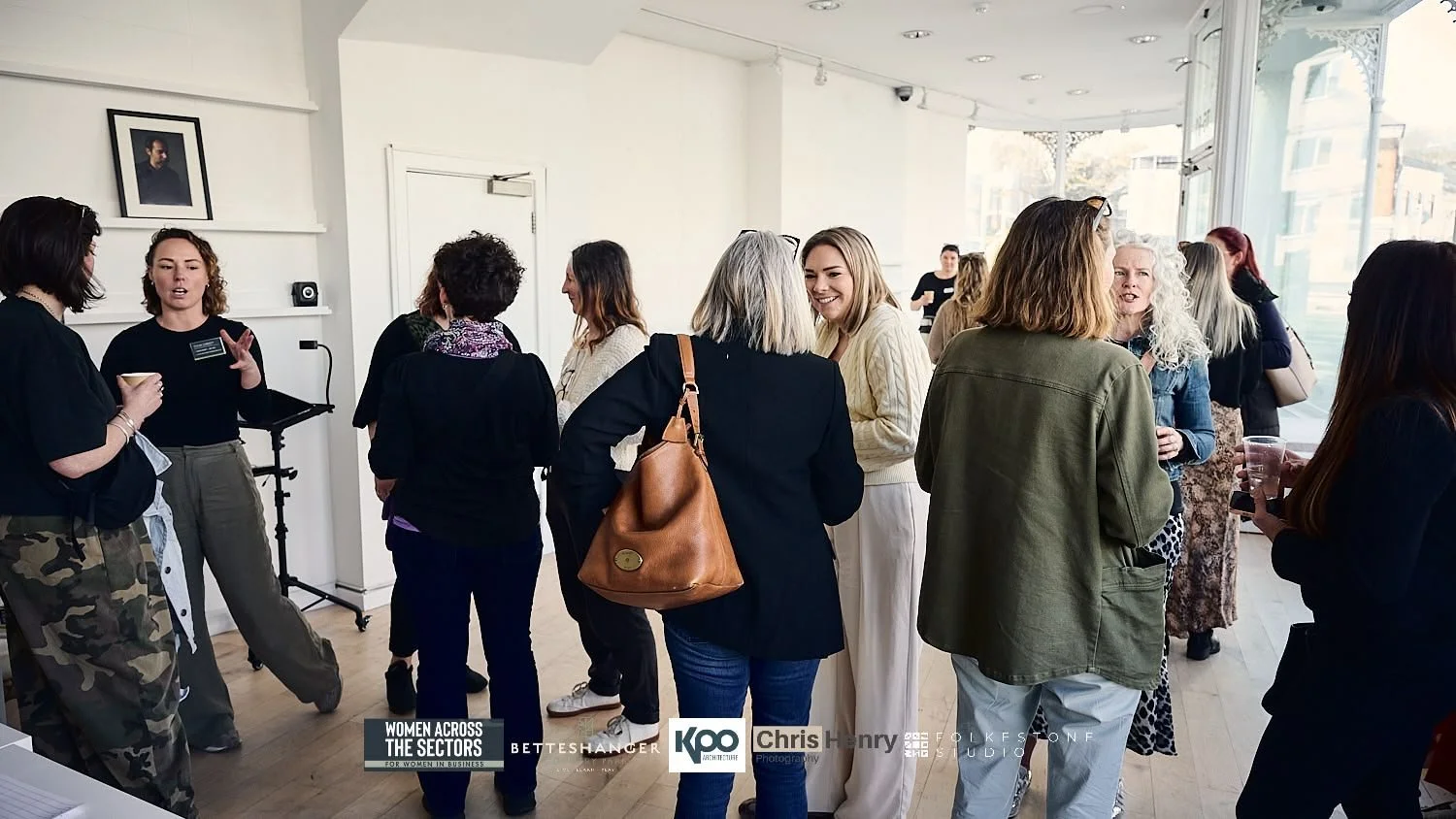 Women gathered in a bright, modern indoor space at a networking or social event, some engaging in conversation, with large windows and framed artwork on the walls.