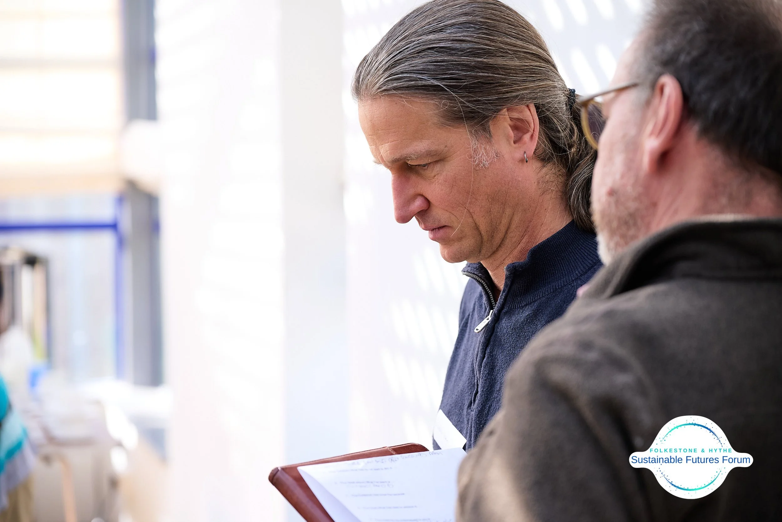 Two men engaged in a serious discussion at an indoor event with a 'Sustainable Futures Forum' logo in the bottom right corner.