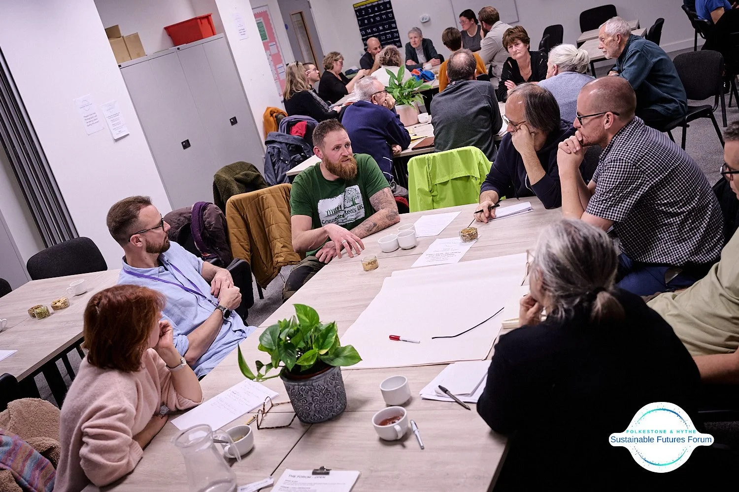 Group of people sitting around a large table in a meeting room, engaged in discussion, with notebooks, papers, cups, and a potted plant on the table.