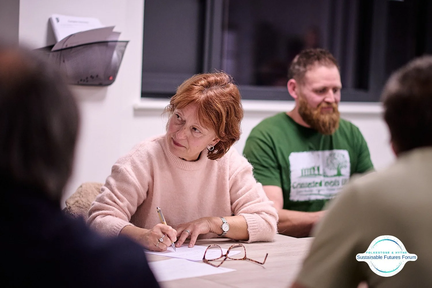 A woman with short red hair, wearing a pink sweater, sitting at a table with papers and reading glasses. She is holding a pen and appears to be engaged in a discussion. There are other people around the table, and a man with a beard in a green t-shir