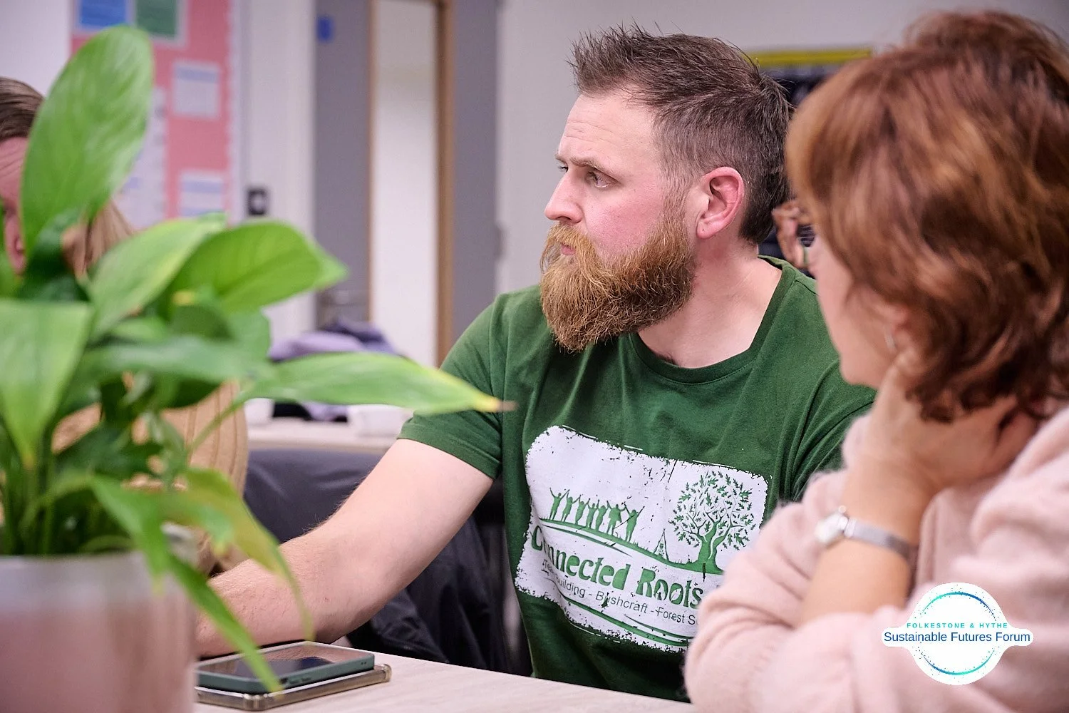 A man with a beard and short hair wearing a green t-shirt is sitting at a table, listening to someone. A woman with red hair wearing a light pink hoodie is sitting nearby, resting her chin on her hand. A green potted plant is in the foreground on the