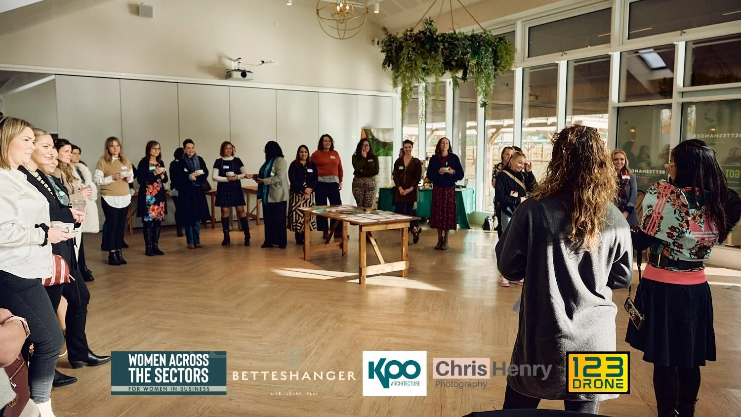 A group of women standing in a semi-circle inside a brightly lit room with large windows, listening to a speaker. There are banners and a table with materials in the background.