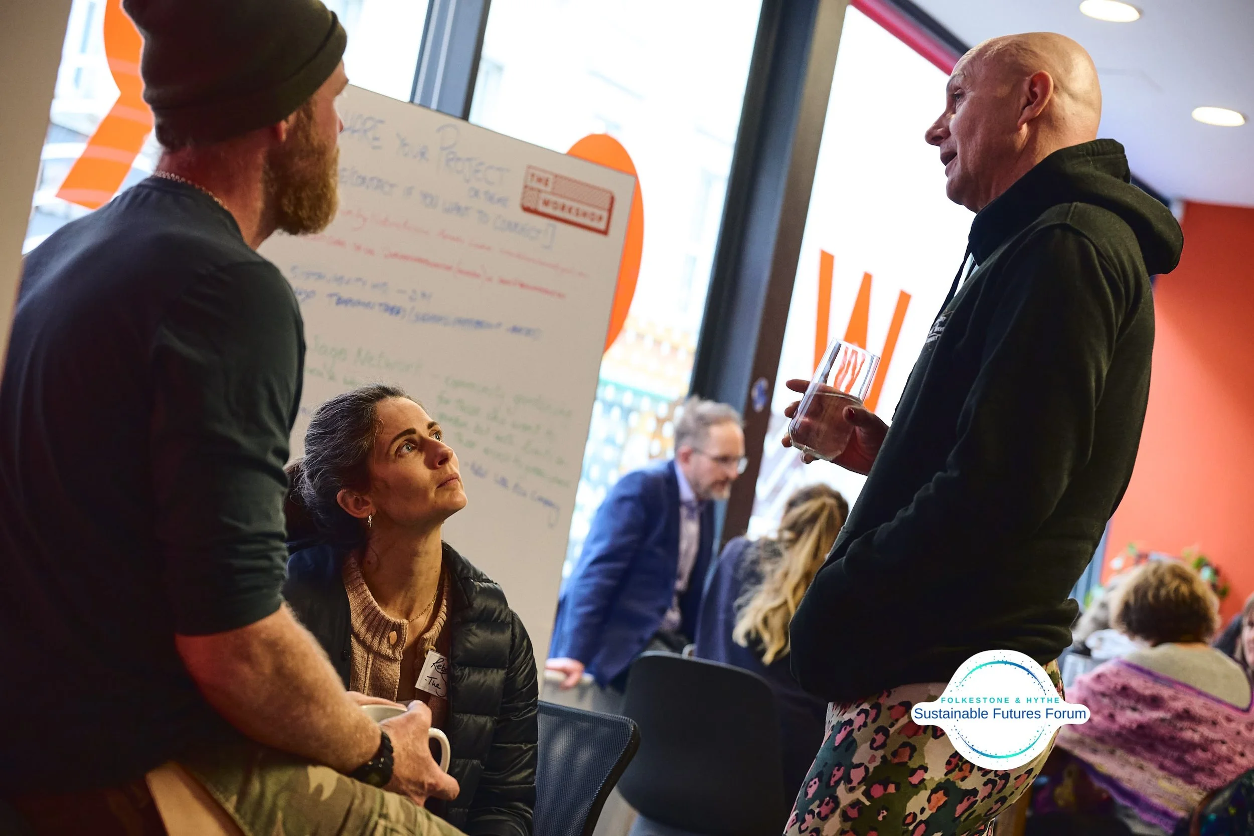 Three people engaged in conversation at a conference or event, with a woman seated and looking up at a man standing, holding a glass, while another man is also present. There is a whiteboard with writing behind them, and other attendees are visible i