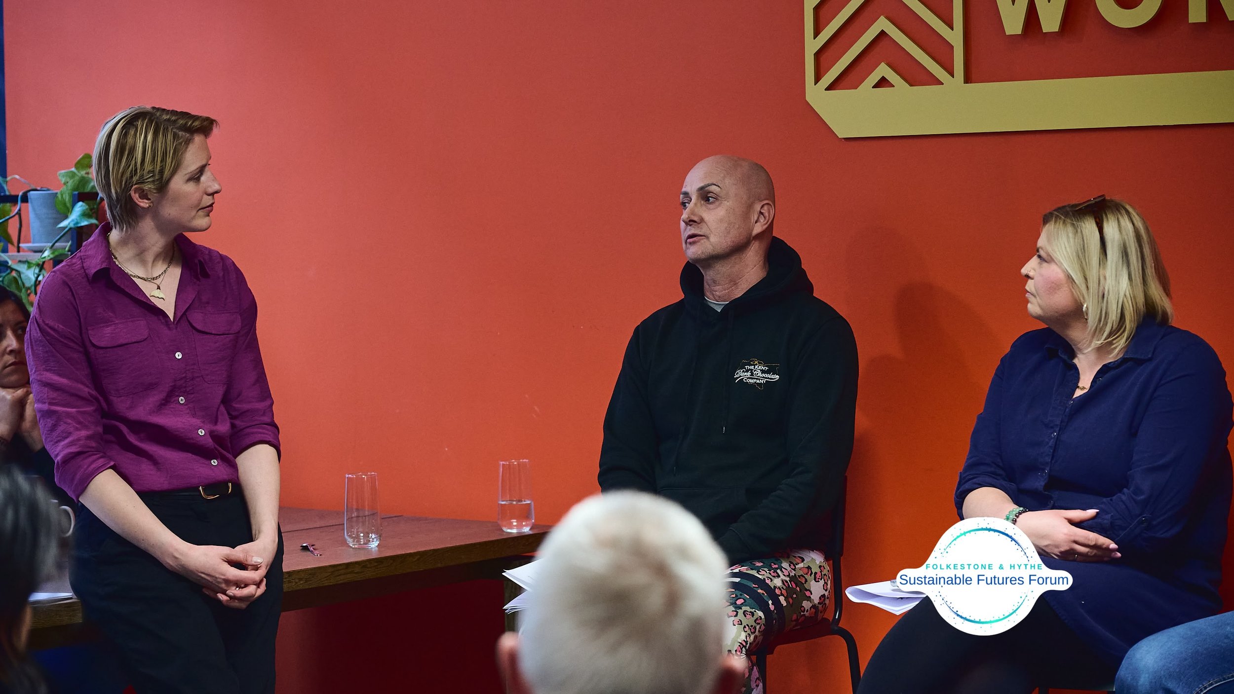 Three people engaged in a conversation at a panel discussion during the Folkestone & Hythe Sustainable Futures Forum, with an orange wall and part of a large sign in the background.