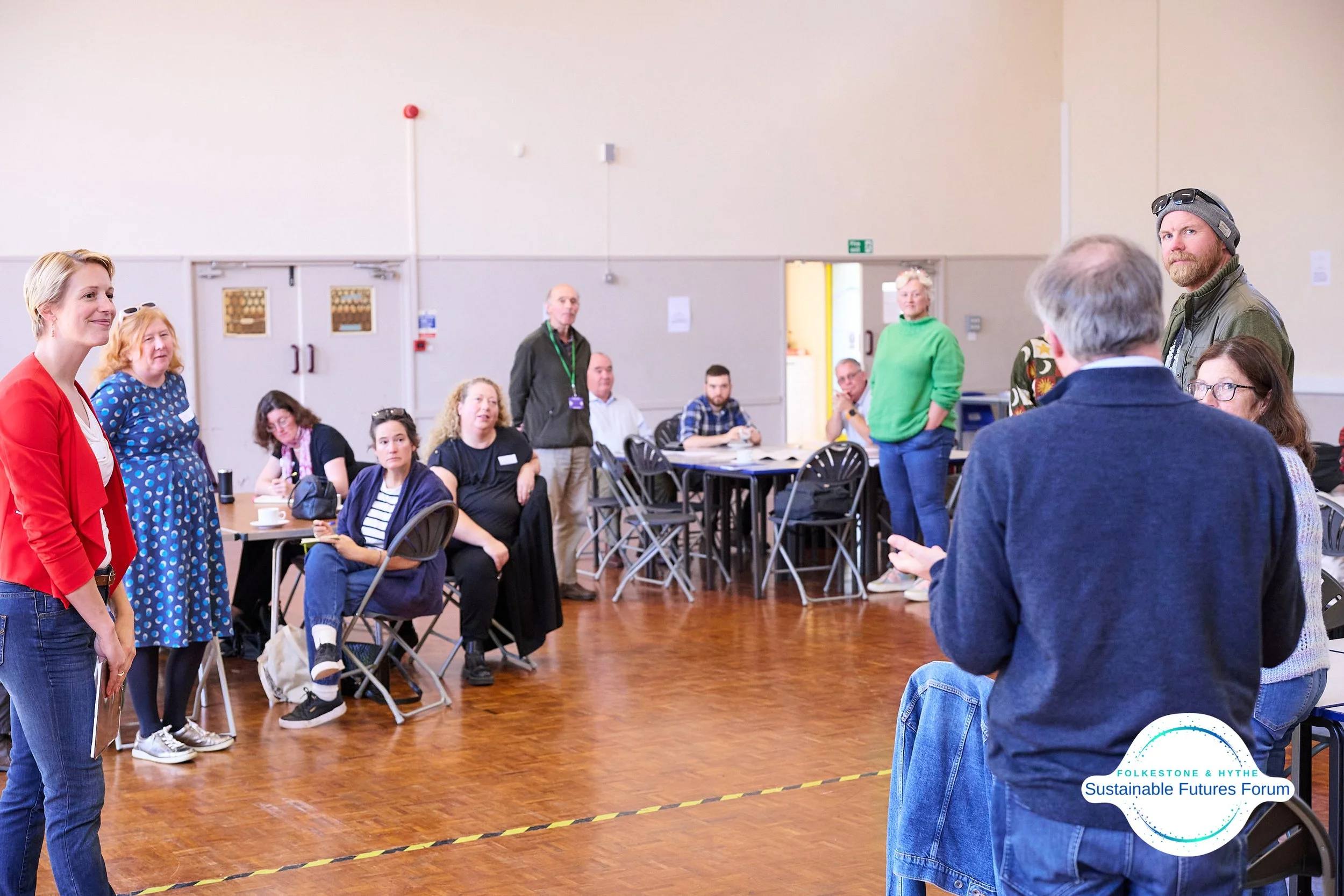 A group of people gathered in a spacious room attending a forum or workshop. Some are seated at tables, others are standing, and a woman in a red blazer is speaking or presenting. There is a logo in the bottom right corner that reads 'FOLKESTONE & HY