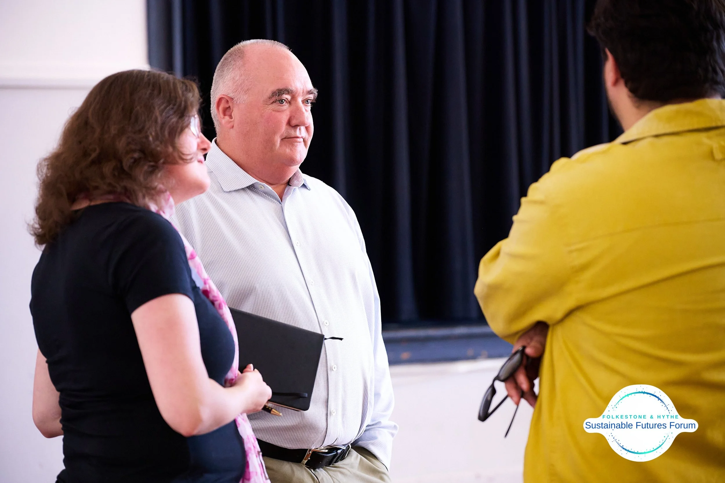 Three people having a conversation at a forum, with a black curtain in the background and a logo for 'Sustainable Futures Forum' in the bottom right corner.