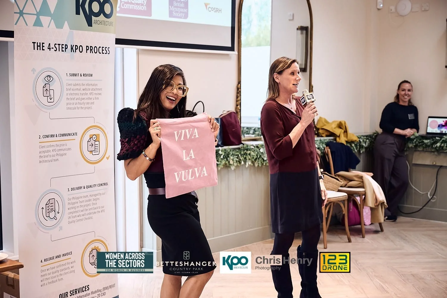 A woman holding a pink cloth with the words "VIVA LA VULVA" written on it, standing next to a woman speaking into a microphone, in a room with a presentation screen and banner about the KPO process. There is another woman standing in the background n