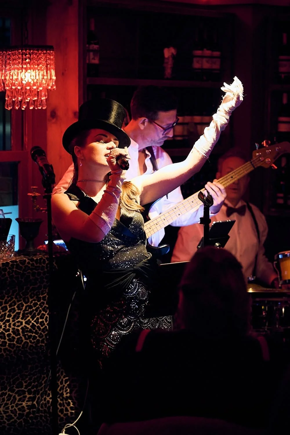 Performer singing into microphone, wearing a top hat and sequined dress, with a band playing in a dimly lit venue with red lighting and wooden decor.