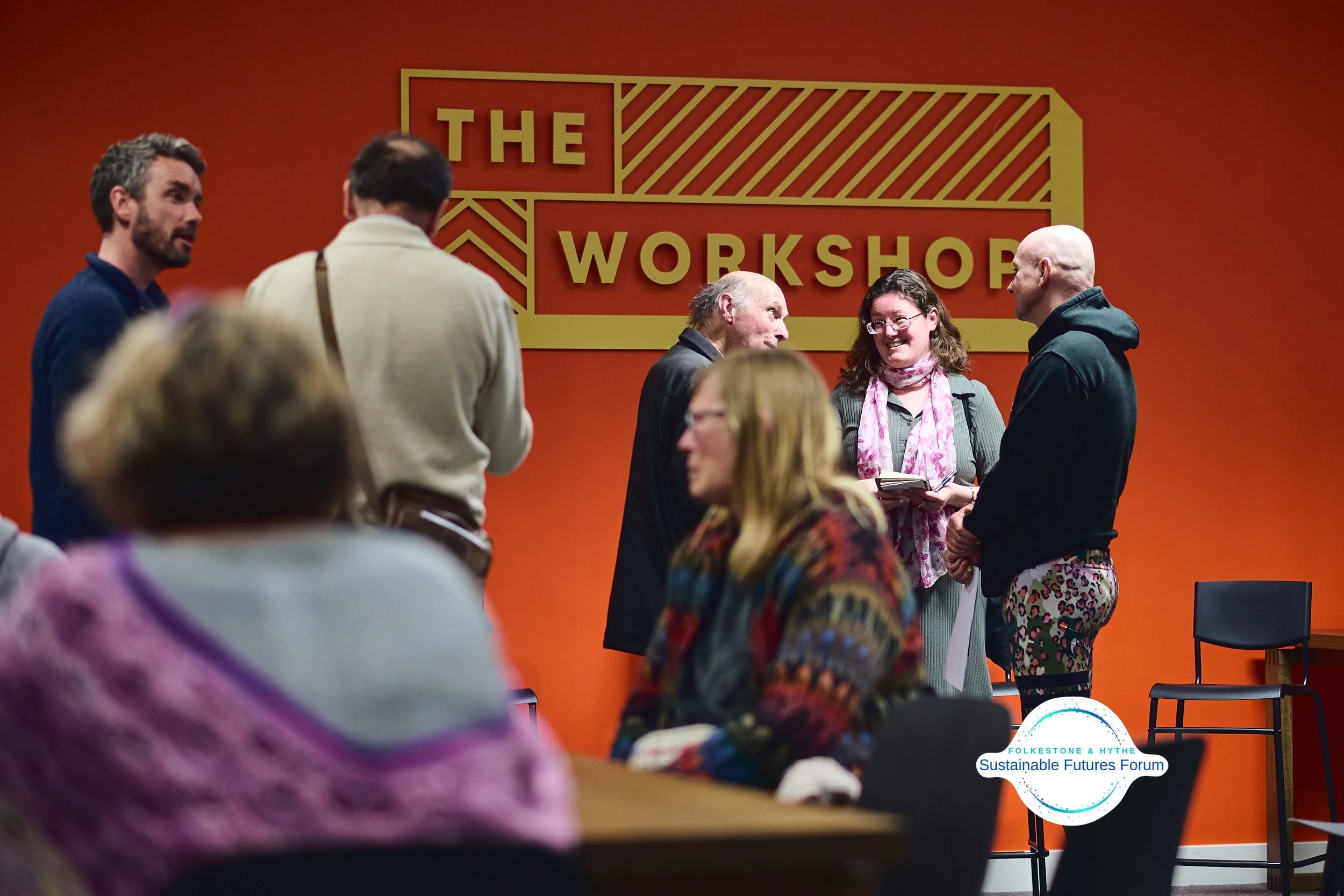 A group of people talking and engaging at a workshop event in a room with orange walls and a large yellow sign that reads 'The Workshop'.