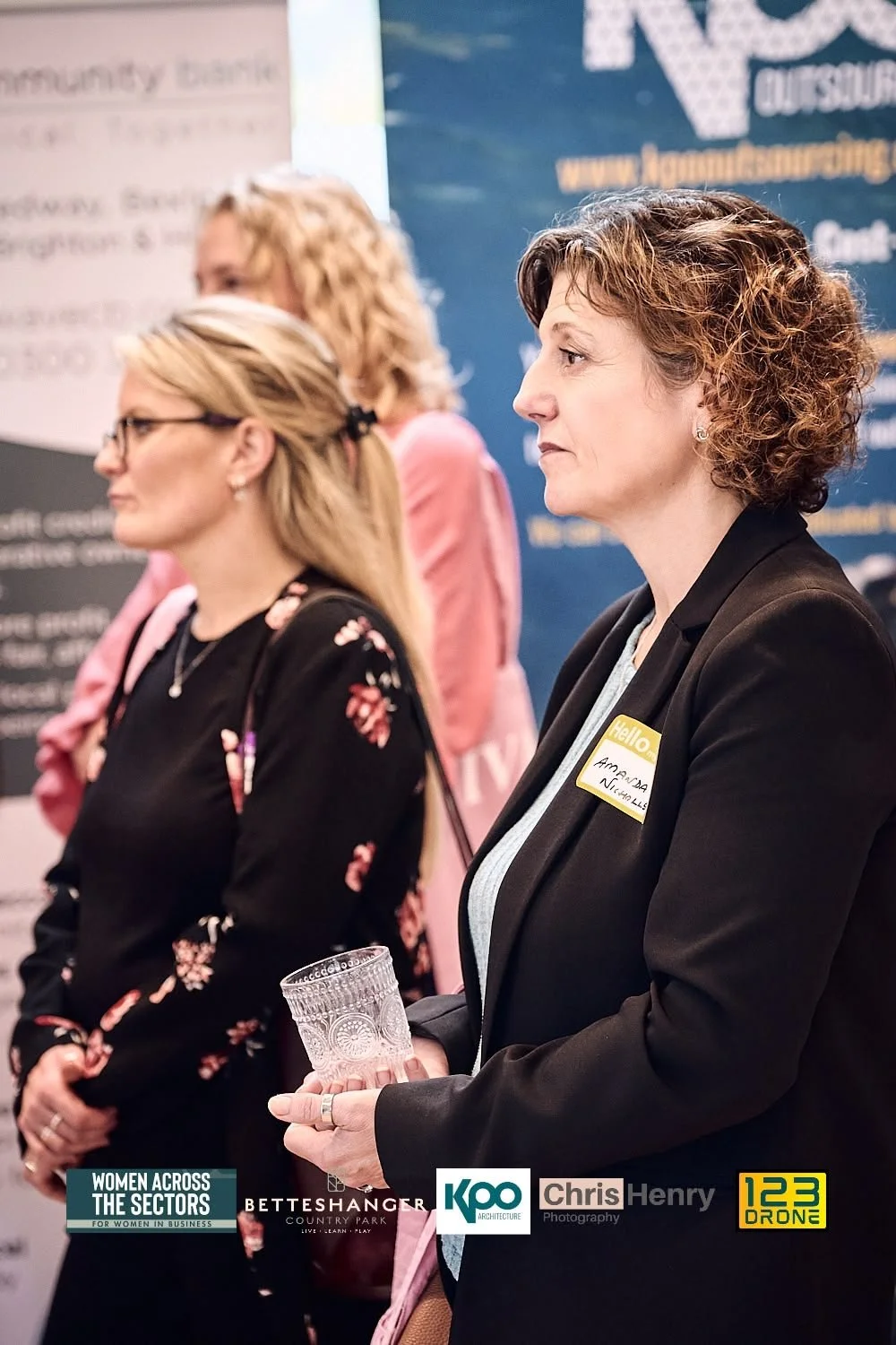 Women attending an event with one woman holding a glass, others with name tags, standing in front of a backdrop with logos and text.