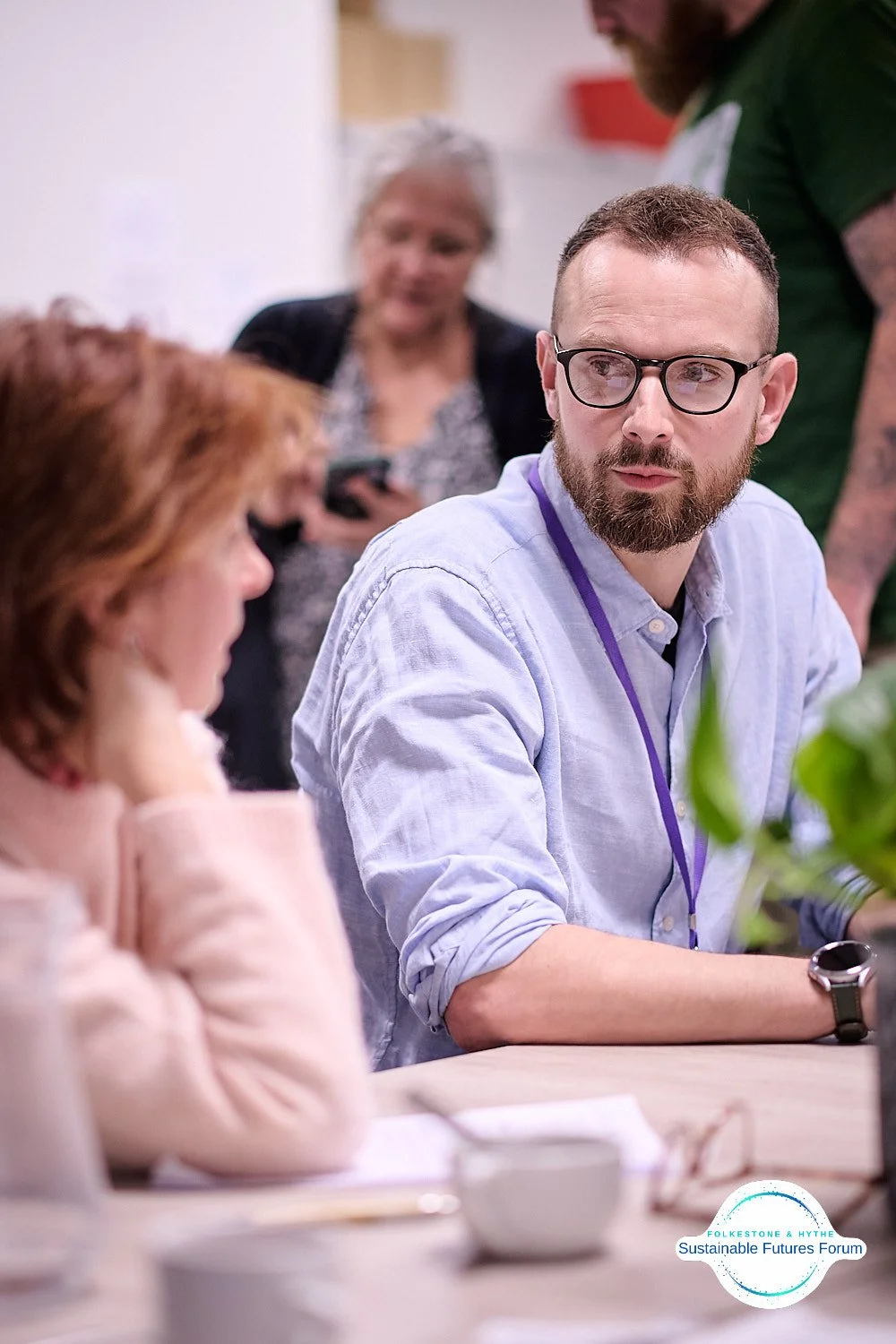 A young man with glasses and a beard sits at a table, talking to a woman with red hair. In the background, a woman with gray hair and a man in a green shirt are engaged in conversation, with the woman looking at her phone.