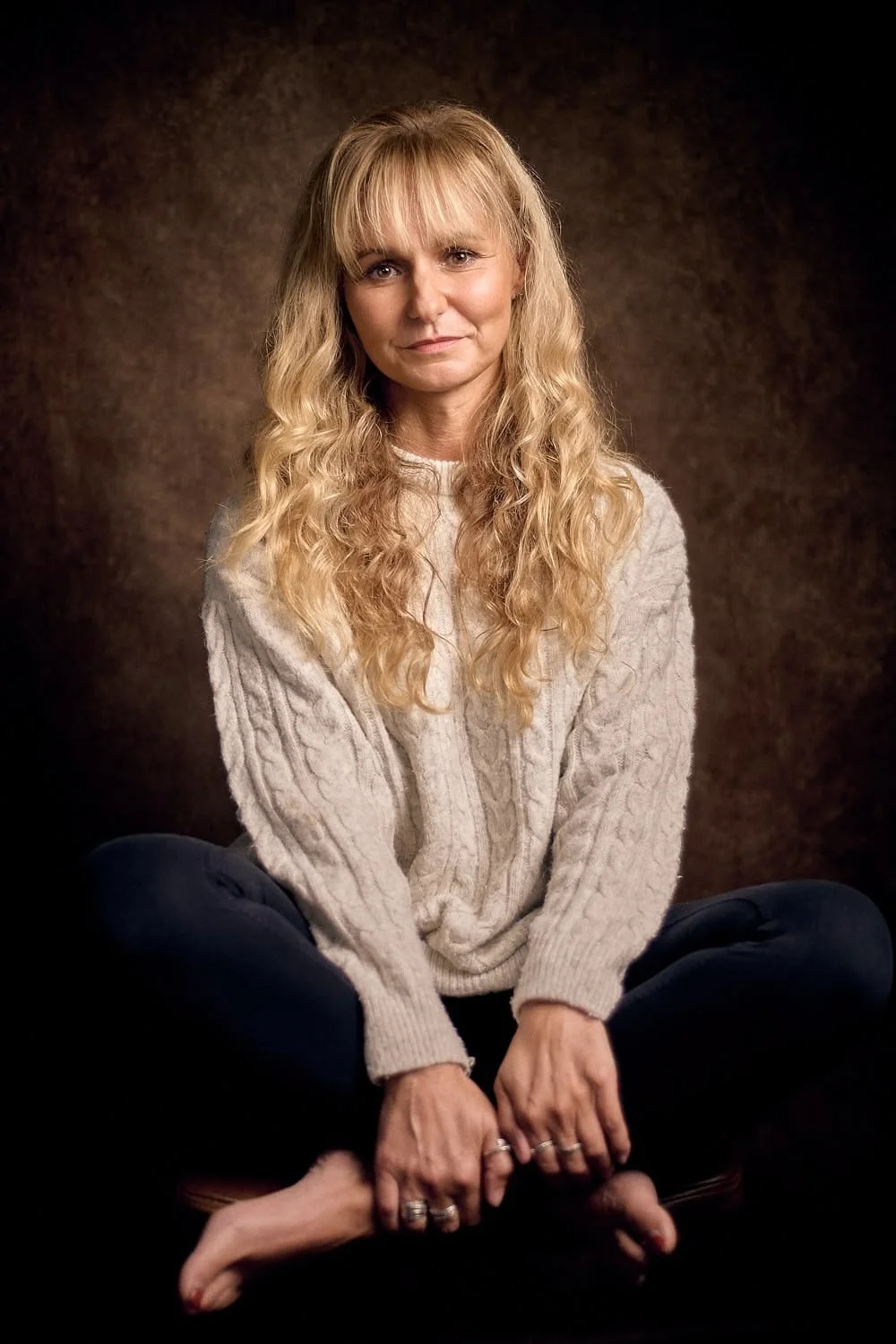 A woman with long, curly blonde hair sitting cross-legged against a brown textured background, wearing a light gray cable-knit sweater and dark pants.