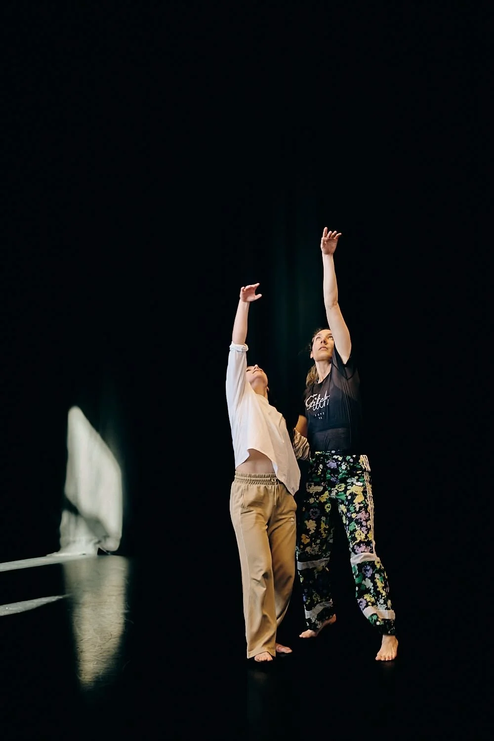 Two women dancing in a dark room, one wearing beige pants and a white top, the other in floral pants and a black top, both reaching upward with their arms.