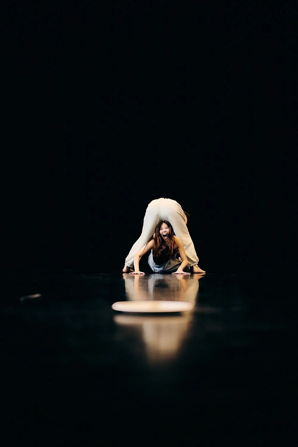 A woman performing a yoga pose on a dark stage with a black background, with another person standing behind her in a backbend, their heads not visible.