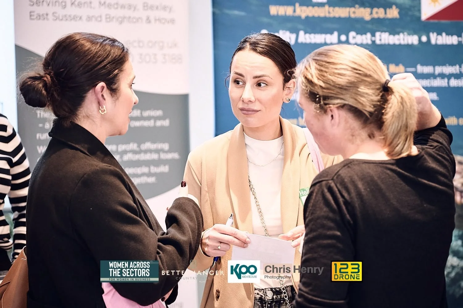 Three women engaged in conversation at a business event with banners and logos in the background.