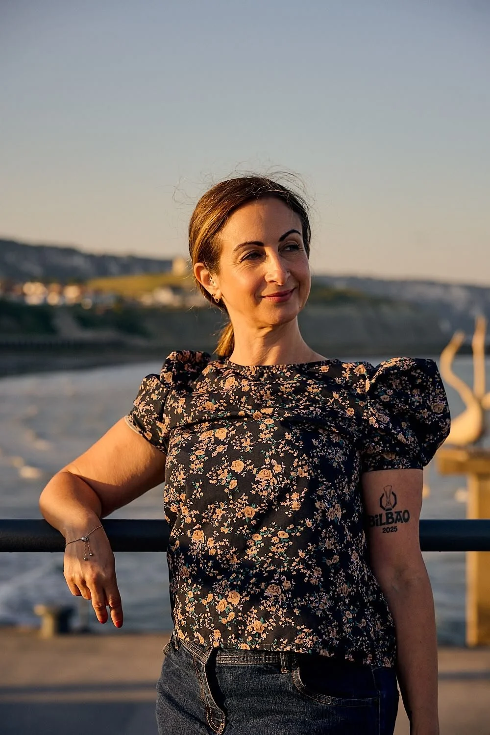 A woman in a floral top standing by a railing outdoors with a river and hills in the background during sunset.
