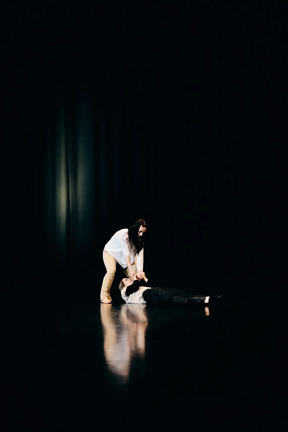 Two women are on a dark stage, one woman is lying on the floor, and the other woman is standing and holding her by the hands in a theatrical or dance performance.