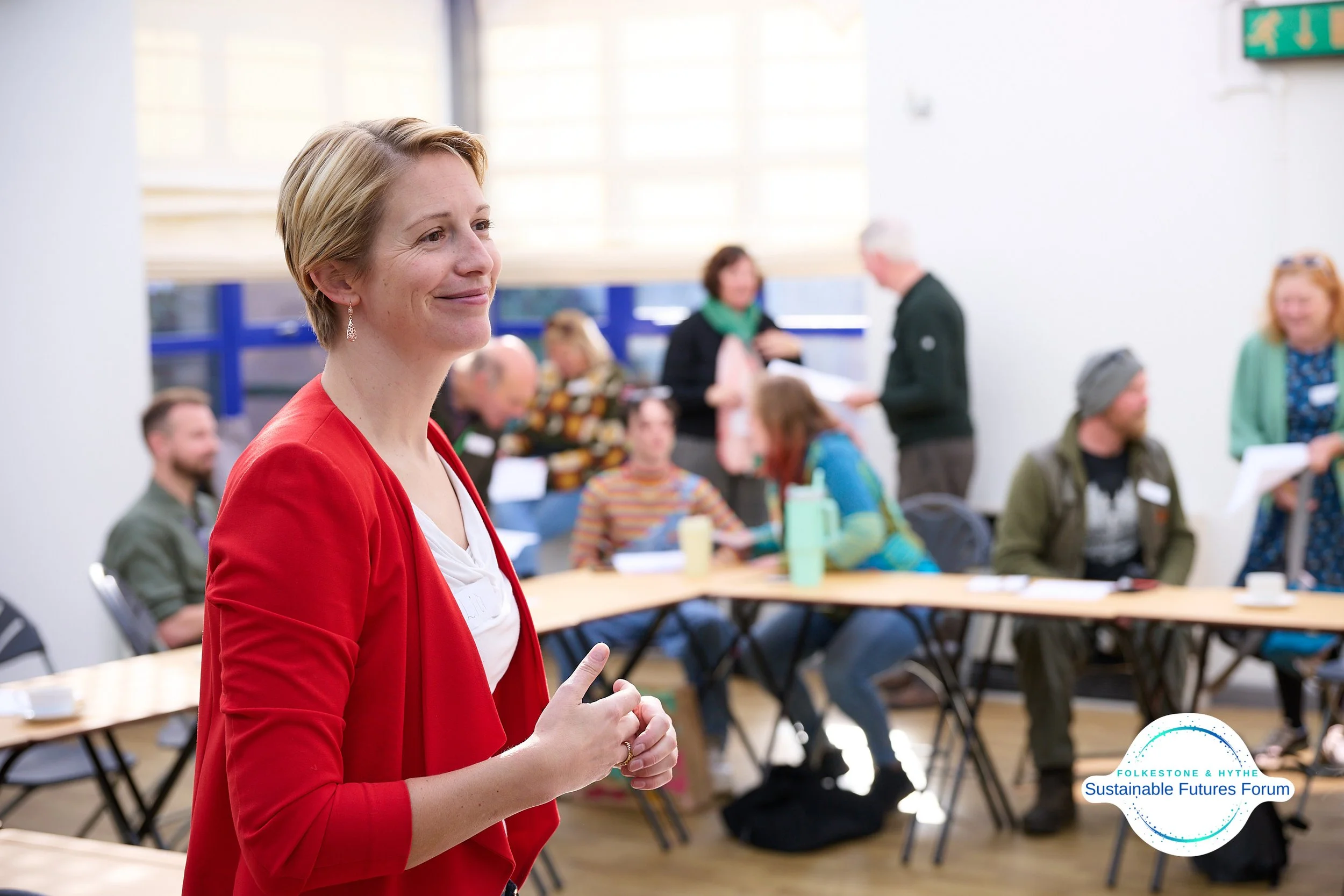 A woman in a red blazer smiling and standing in a conference room with a group of people seated at tables in the background.