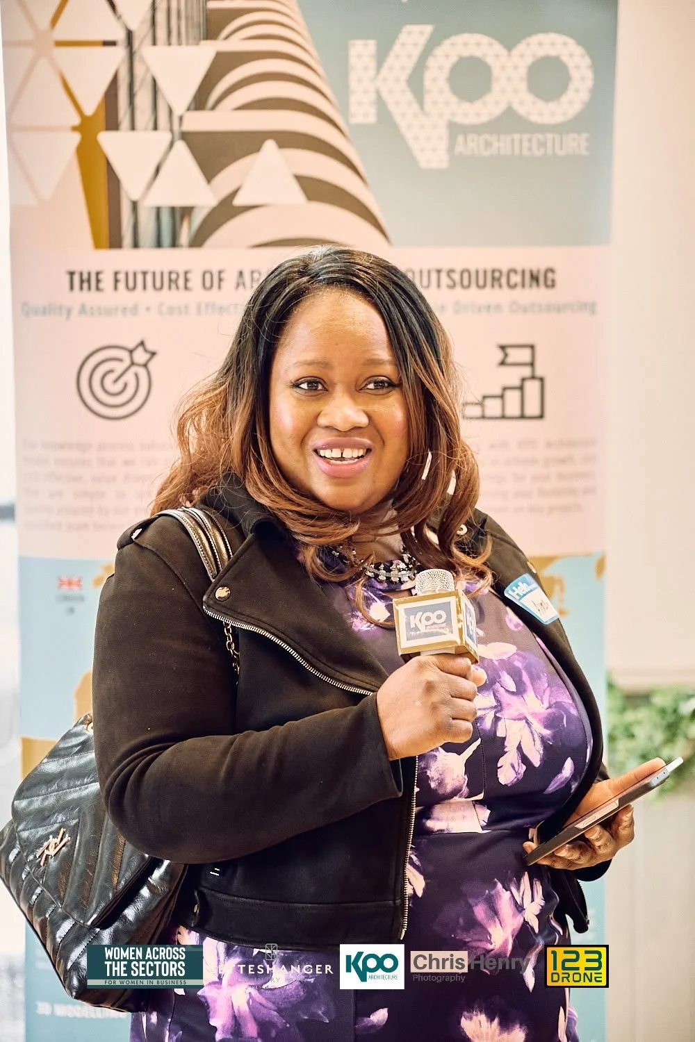 A woman holding a microphone with a "KOO Architecture" logo, standing in front of a fabric banner promoting architecture outsourcing and holding a smartphone.