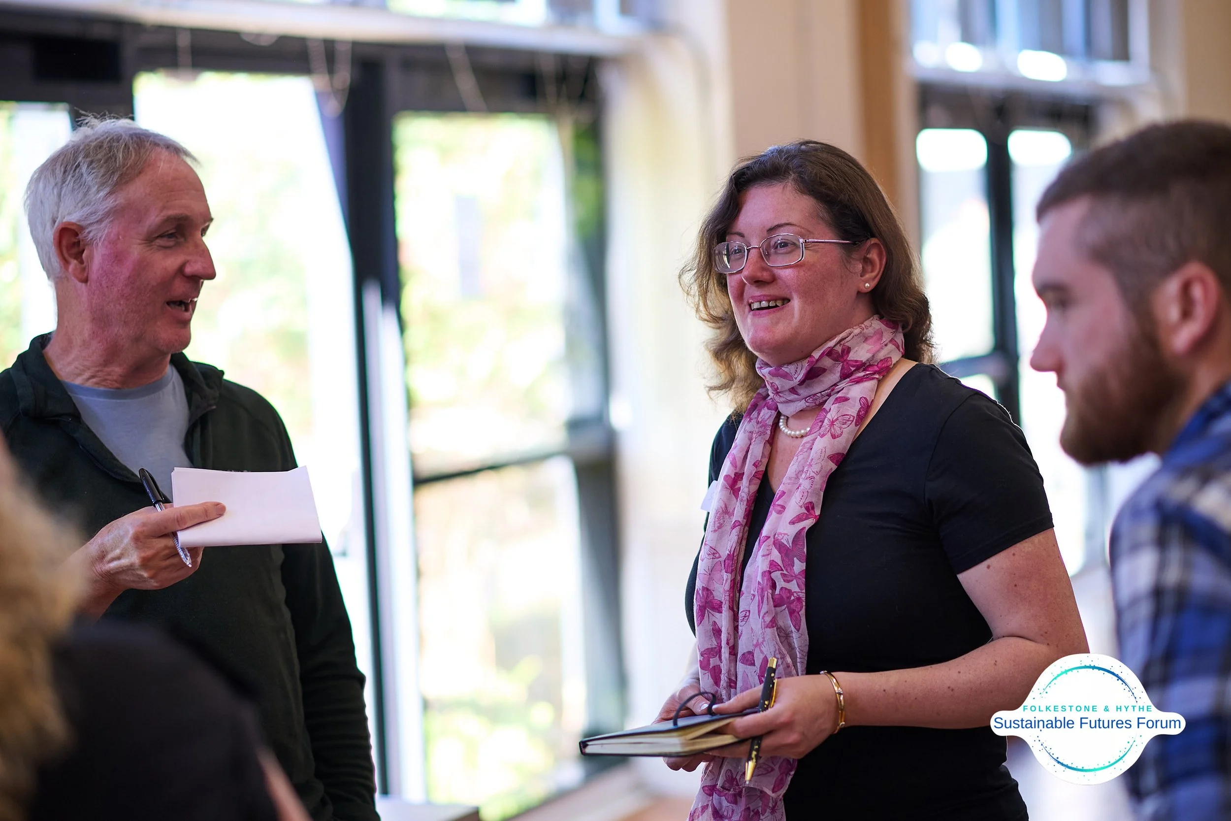 People smiling and talking at a community event or forum, with windows in the background and a logo that says 'Folkestone & Hythe Sustainable Futures Forum' in the bottom right corner.