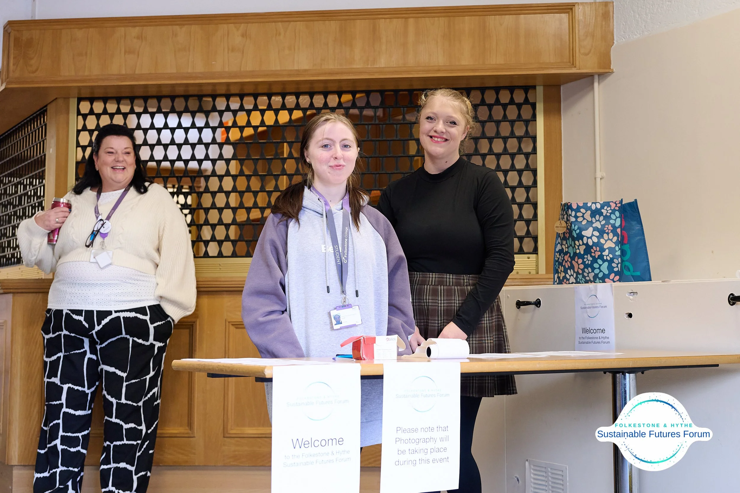 Three women standing behind a registration table at the Folketone & Hythe Sustainable Futures Forum, with one woman on the left holding a drink and smiling, and two women in front of the table looking at the camera.