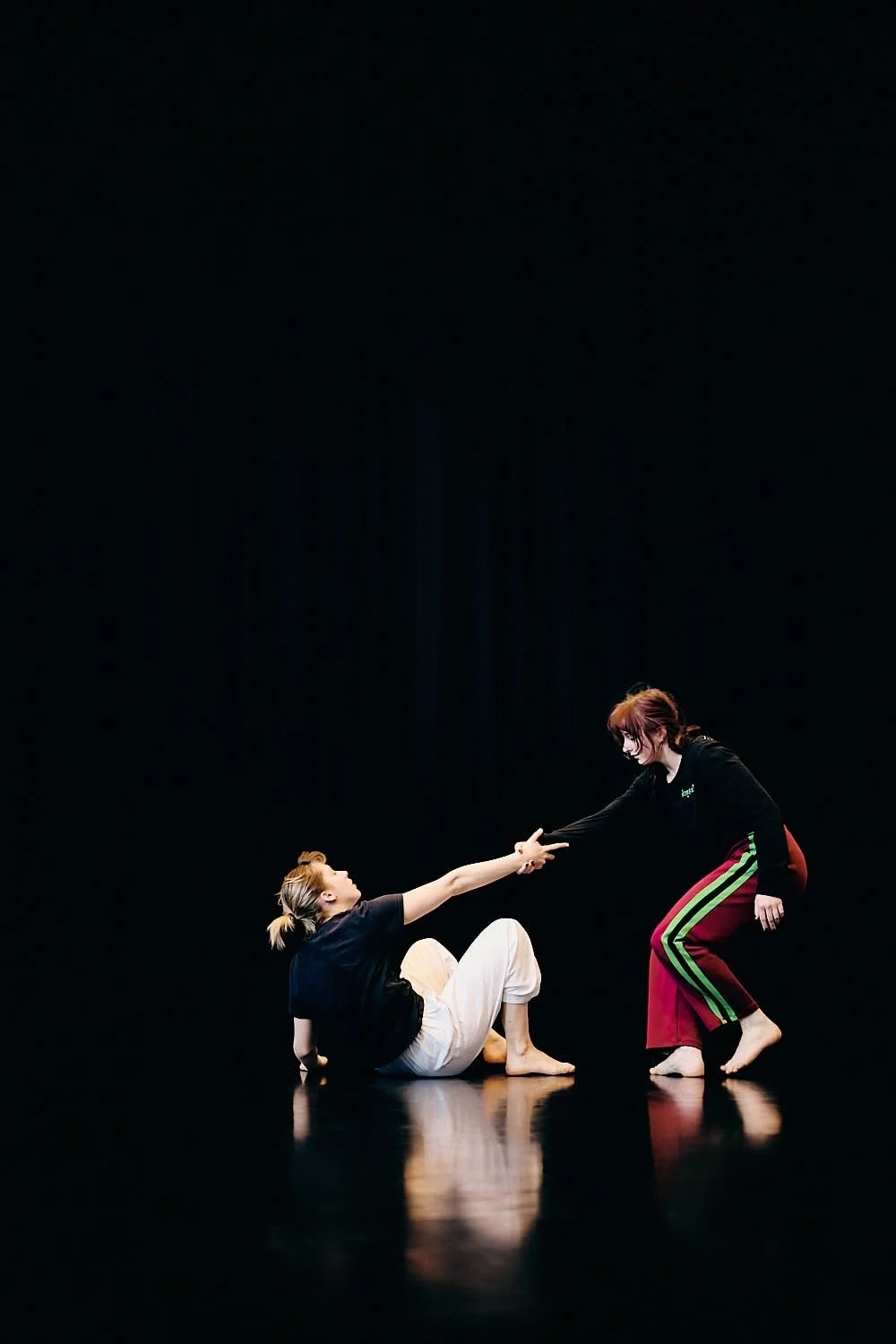 Two women practicing dance or theater on a dark stage, one sitting on the floor reaching out with her hand, the other kneeling and holding her hand.