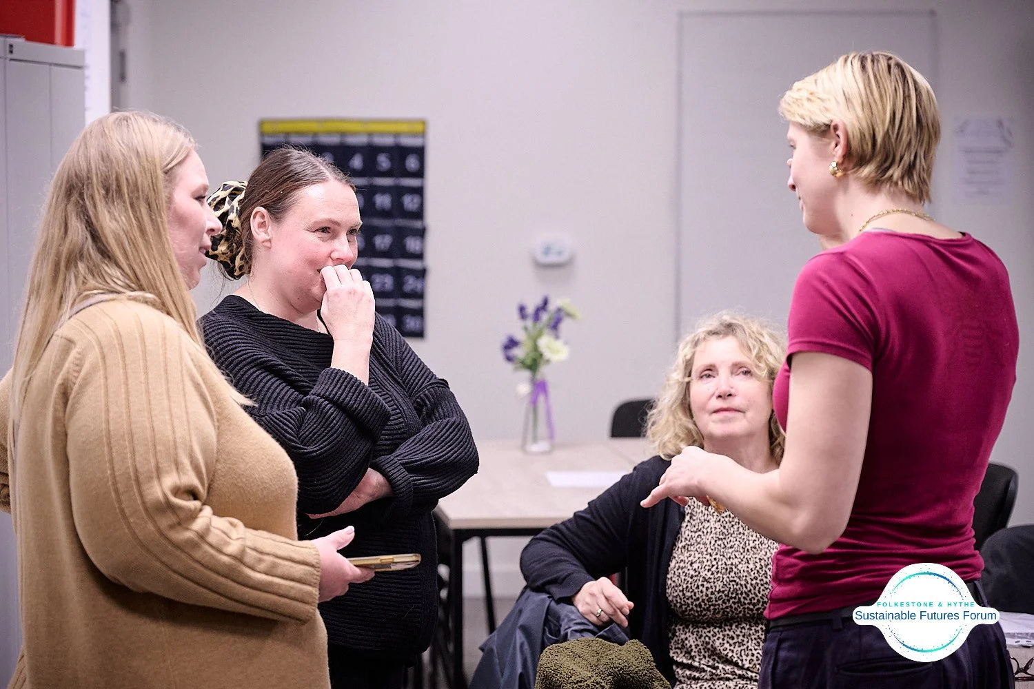 Four women having a serious conversation in an indoor setting, with balloons and a flower arrangement in the background.