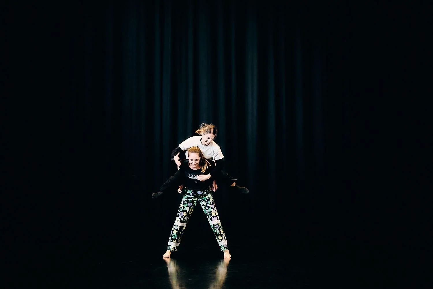 Two women performing an acrobatic dance move on stage, one lifting the other, against a dark backdrop with stage curtains.