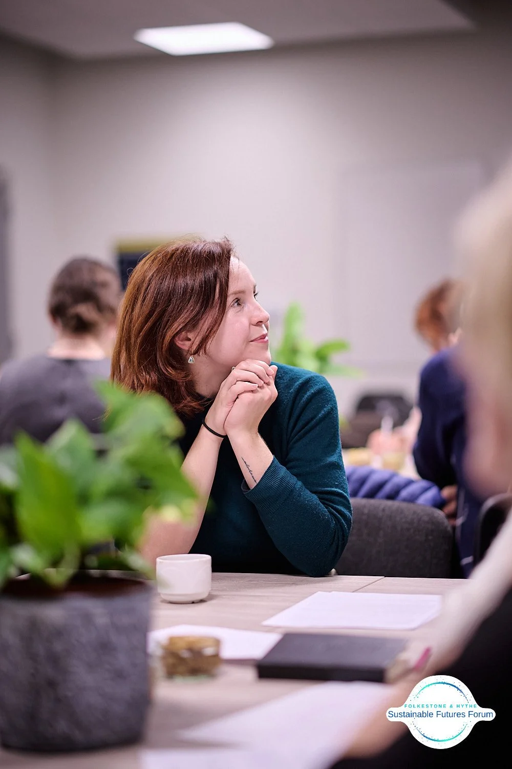 A woman with reddish-brown hair sitting at a table, looking attentively to the side with her hands clasped under her chin during a meeting or discussion. There are plants, papers, and a cup on the table with a logo for the Folketone & Ayth Sustainabl