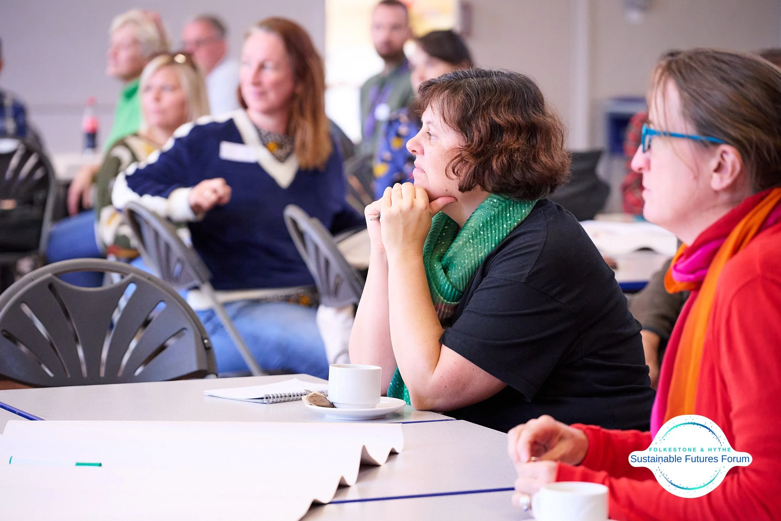 Group of people sitting at a conference table listening attentively, with some taking notes and sipping coffee, during the Folkestone & Hythe Sustainable Futures Forum