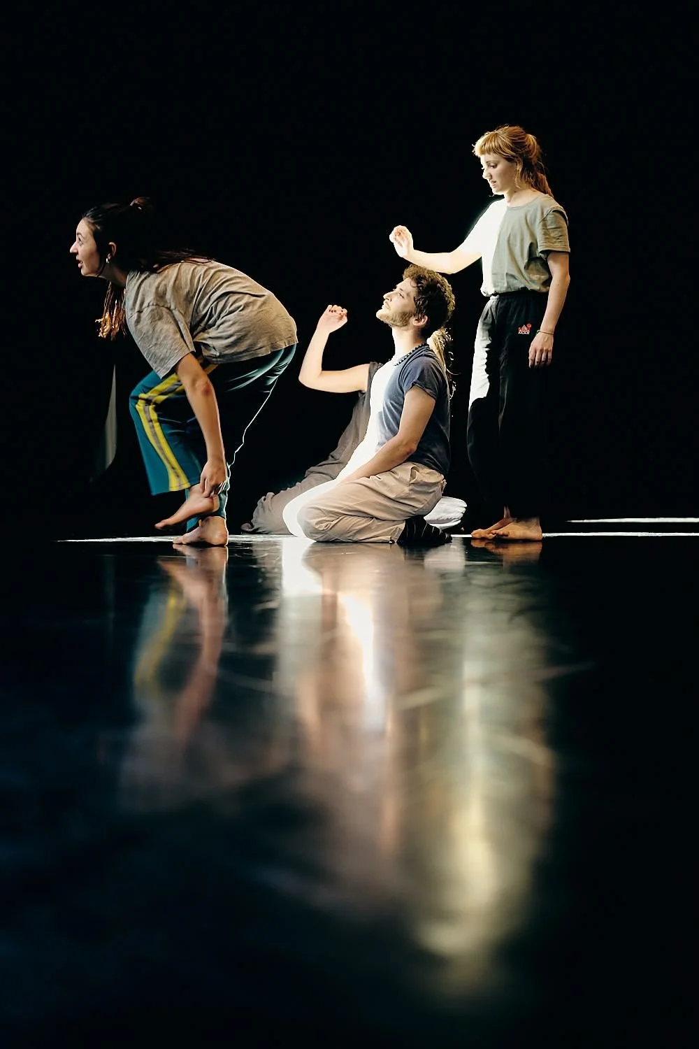 Four young people performing a theatrical scene on a dark stage, with some sitting and others standing, dressed casually, illuminated by stage lighting.