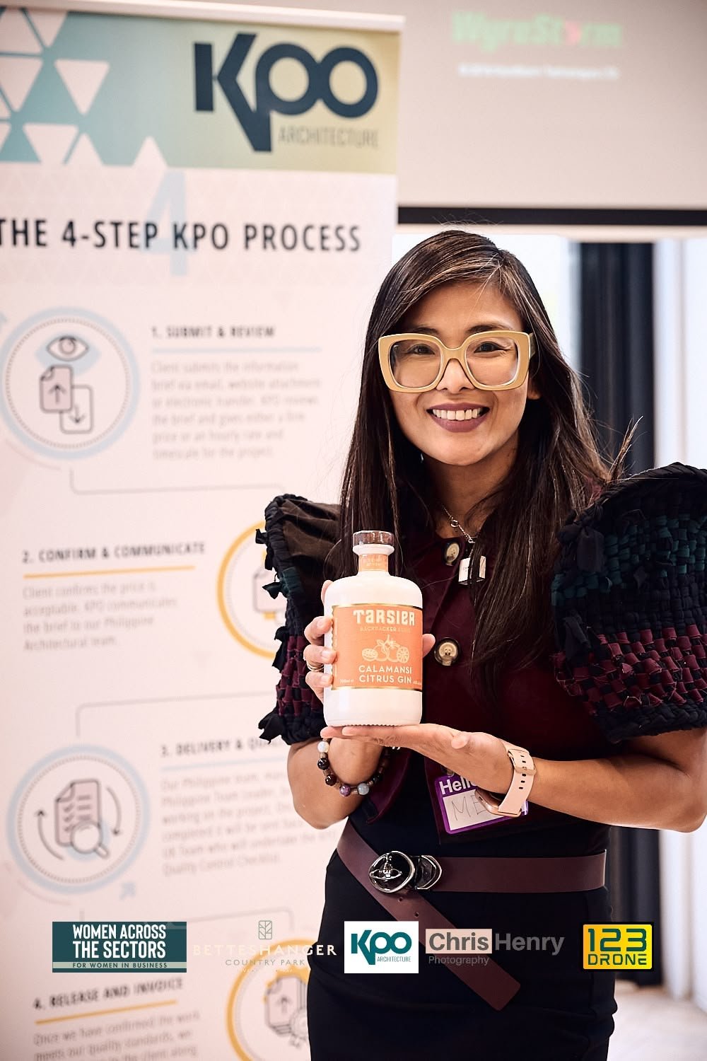 A woman with long dark hair, wearing large yellow glasses and a black outfit, holding a bottle of calamansi citrus gin, smiling at a conference or event with a KOO architecture poster in the background.