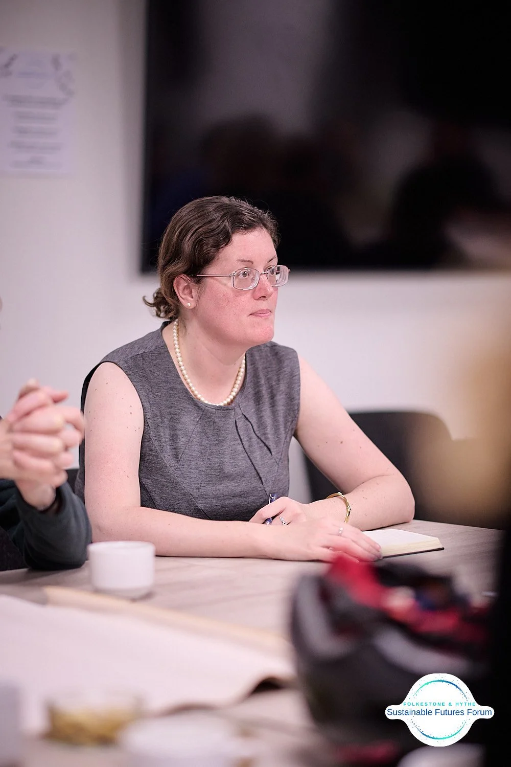 A woman with short curly brown hair, glasses, and wearing a gray sleeveless dress and a pearl necklace, sitting at a conference table with a notepad, a cup, and other items in front of her, listening attentively.