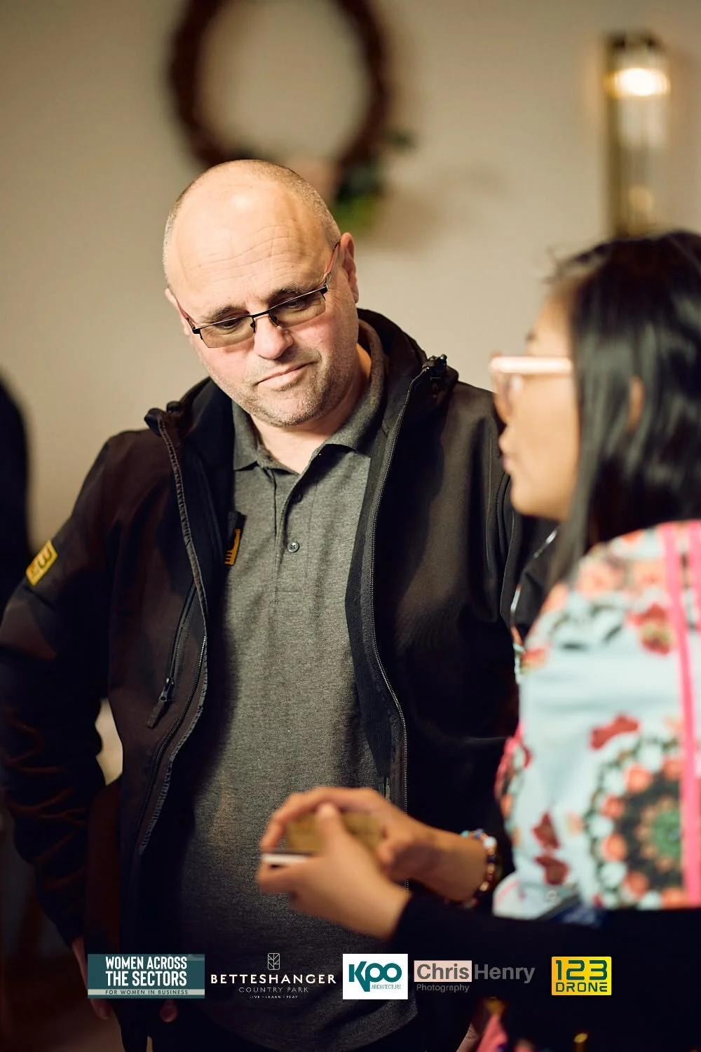 A man with glasses and a bald head is looking at a woman who is holding a phone. The woman has dark hair and is wearing glasses and a floral-patterned top. The background includes a mirror and a wall light, suggesting they are indoors.