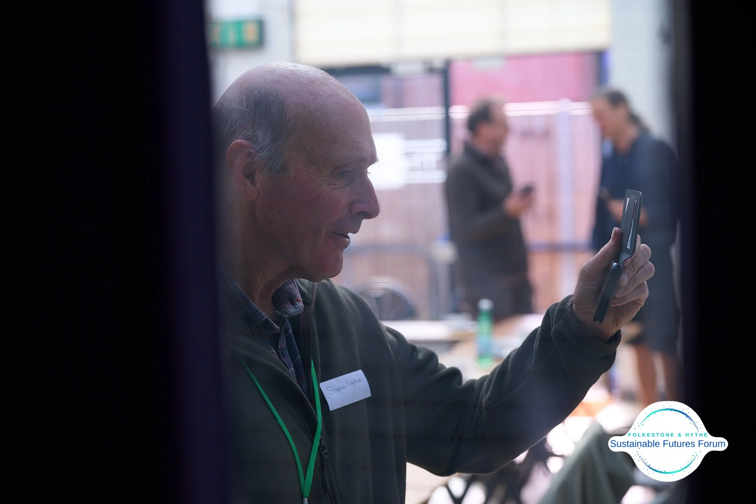 An elderly man with a bald head and gray hair on the sides, wearing a dark jacket and a name tag, is looking at and holding a flip phone. Two women in the background are engaged in conversation and looking at their phones. The setting appears to be a