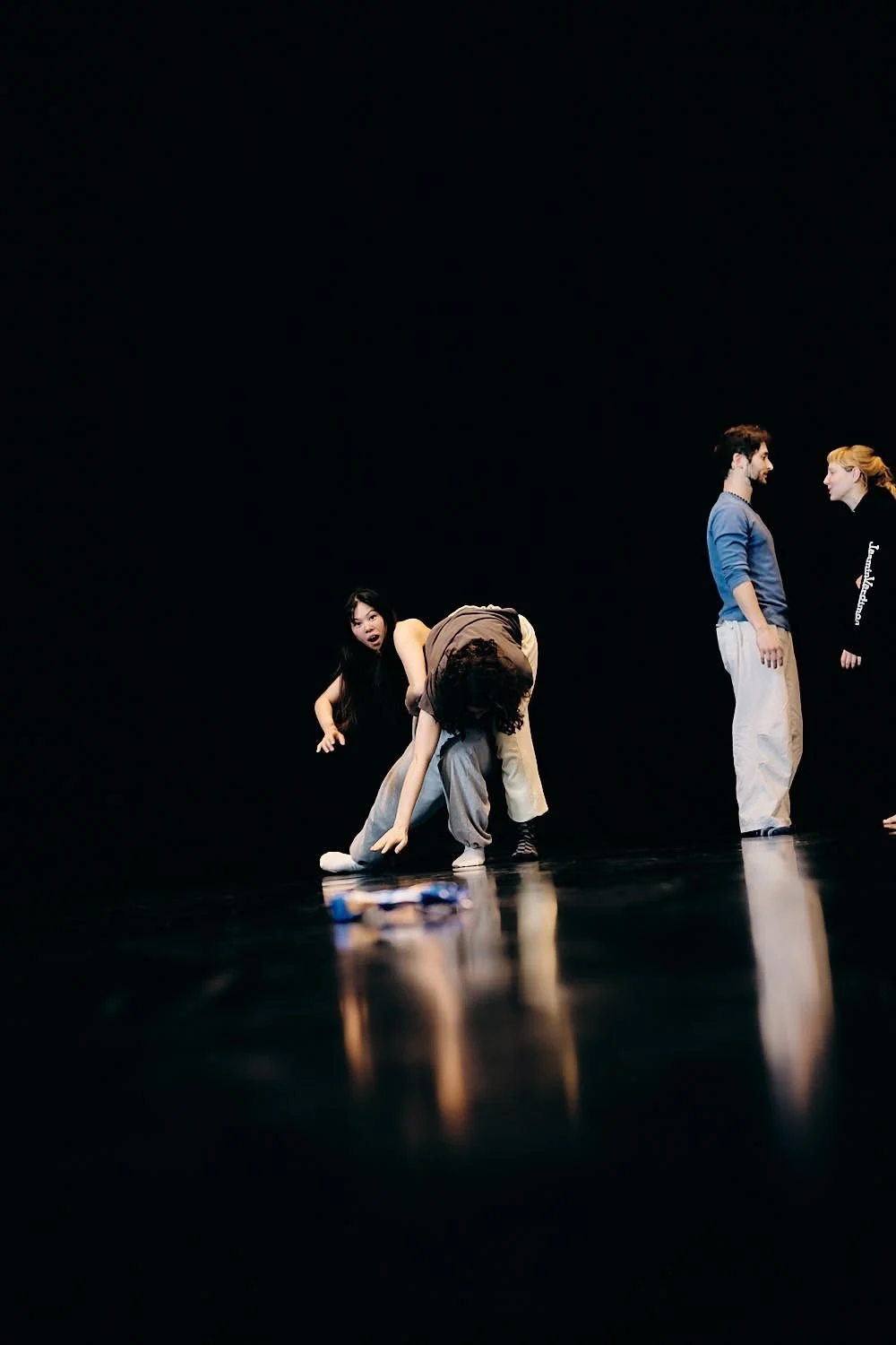 Four people on stage against a black background, with one person bending down, two standing and talking, and one crouching, all in casual clothing.