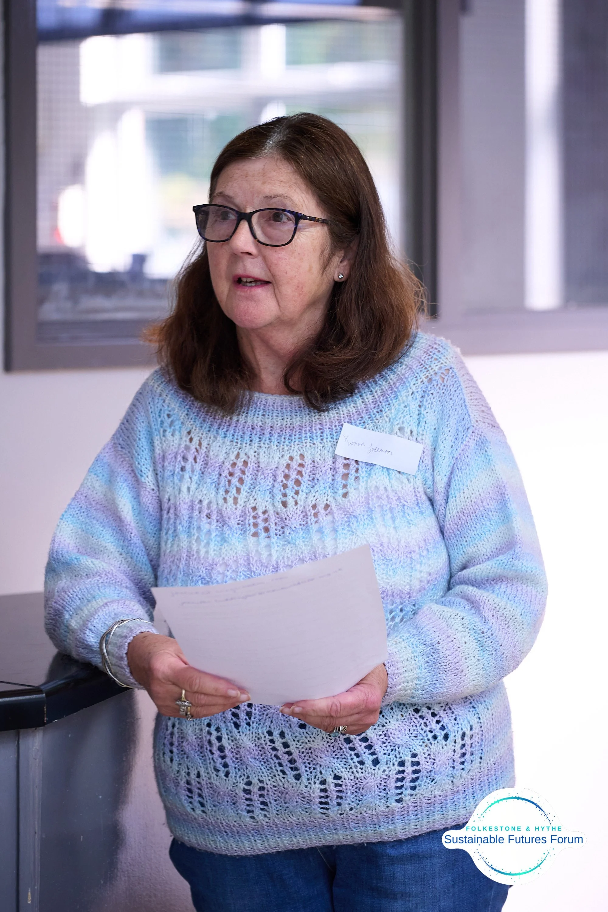 A middle-aged woman with brown hair and glasses speaking at a forum, wearing a pastel-colored knitted sweater and holding a paper, with a label that says her name, in a room with windows and a logo in the bottom right corner reading 'Folkestone & Hyt