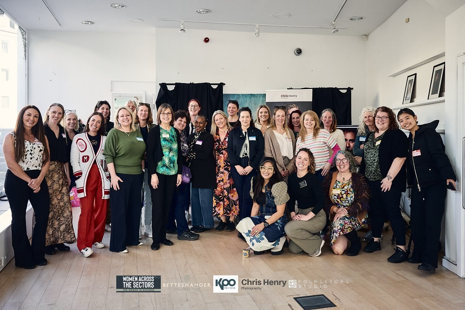 Group photo of diverse women gathering indoors, some standing, some kneeling, smiling for the camera, with a white wall, monitor, and framed pictures in the background.