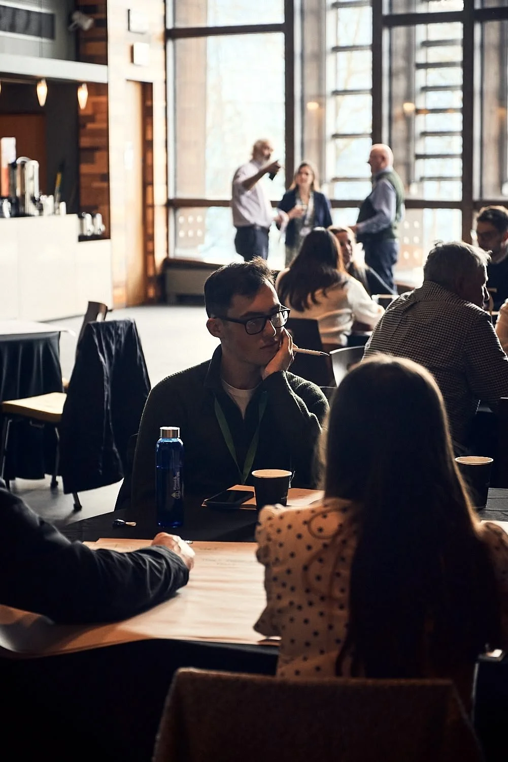 People sitting at tables in a modern cafe or conference space with large windows, while others are standing and talking in the background.