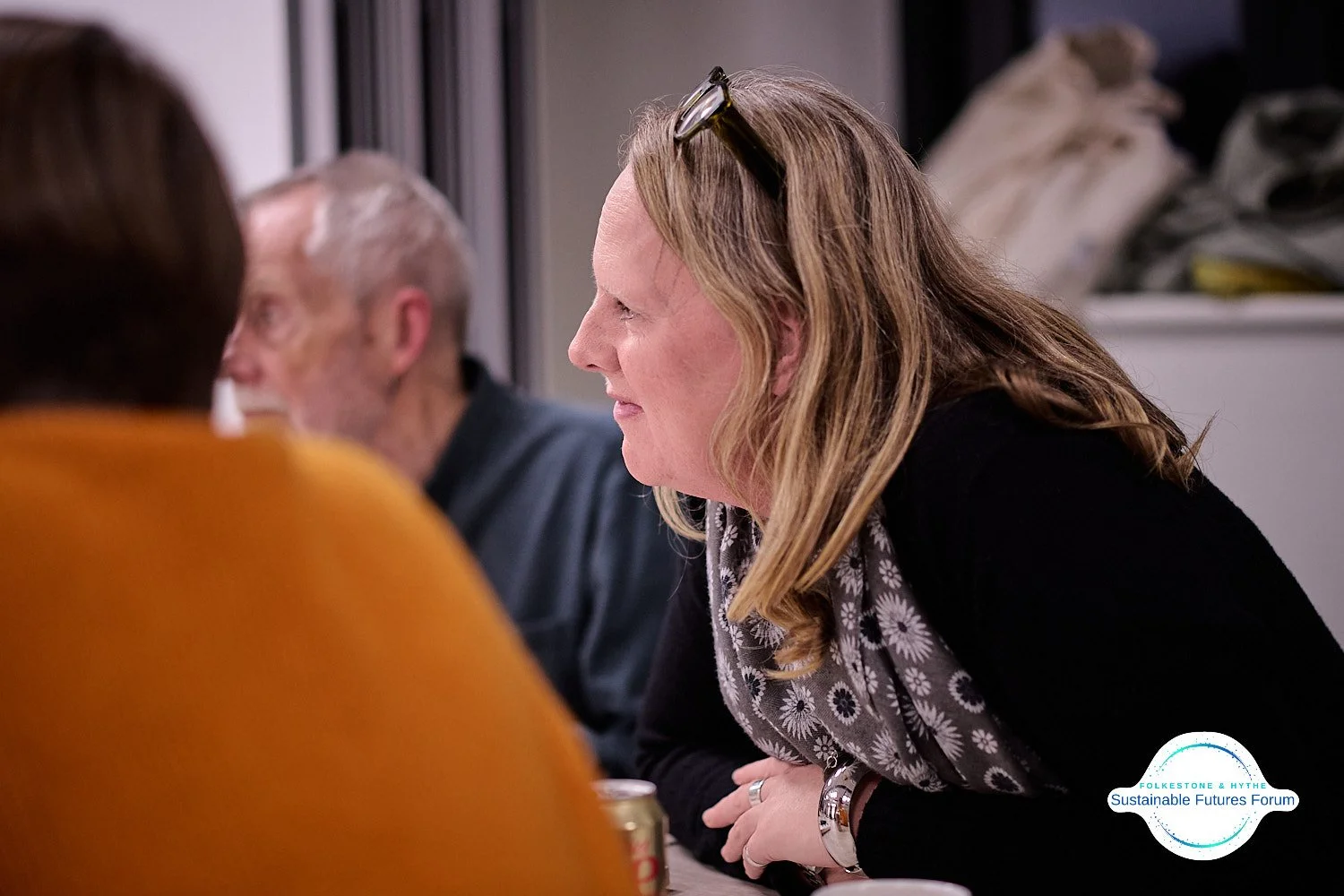 A woman with blonde hair, glasses on her head, and a gray patterned scarf, sits at a table engaged in conversation, with two men seated nearby, in an indoor setting.