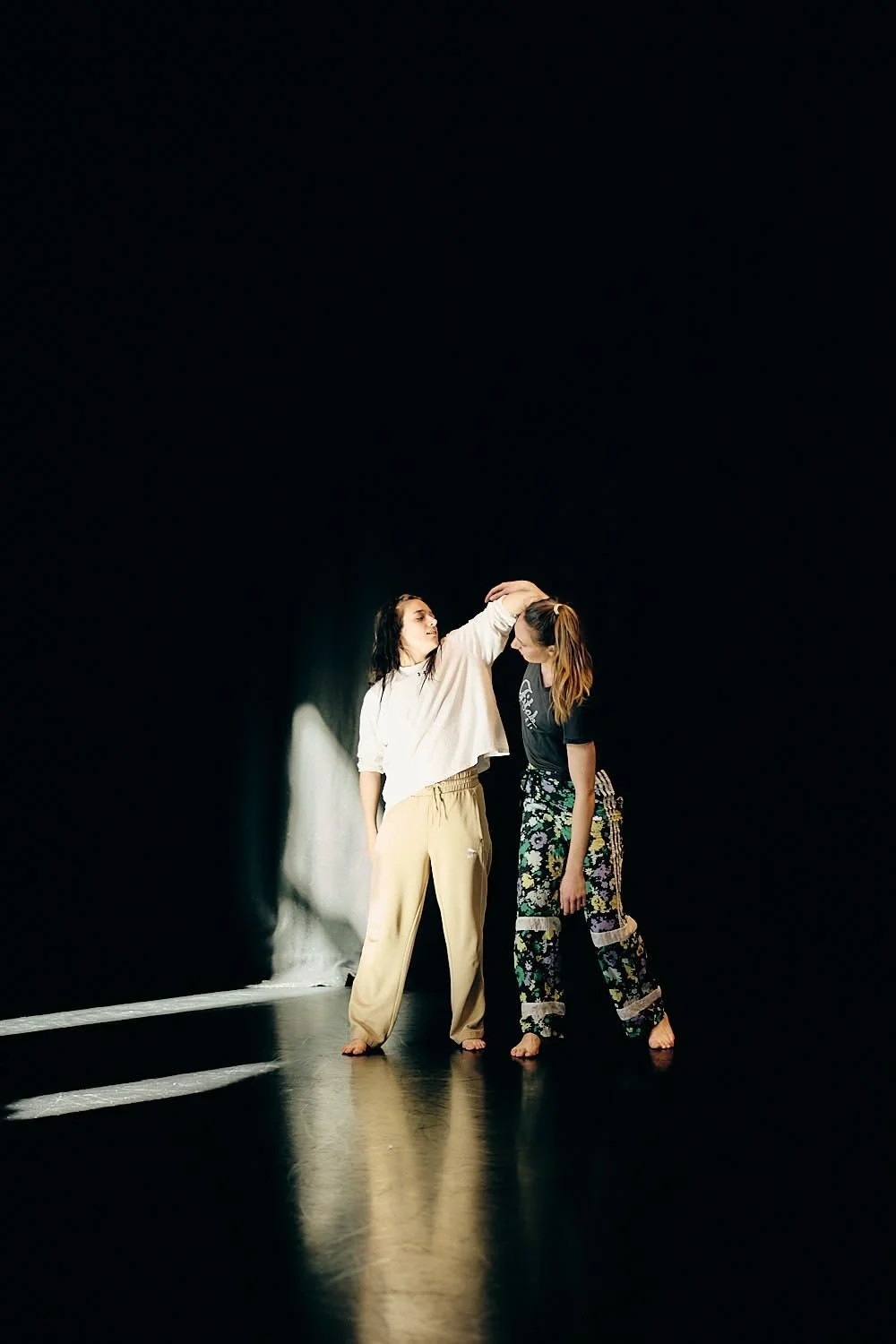 Two women dancing on a dark stage with minimal lighting, one is touching the other's head, both barefoot.