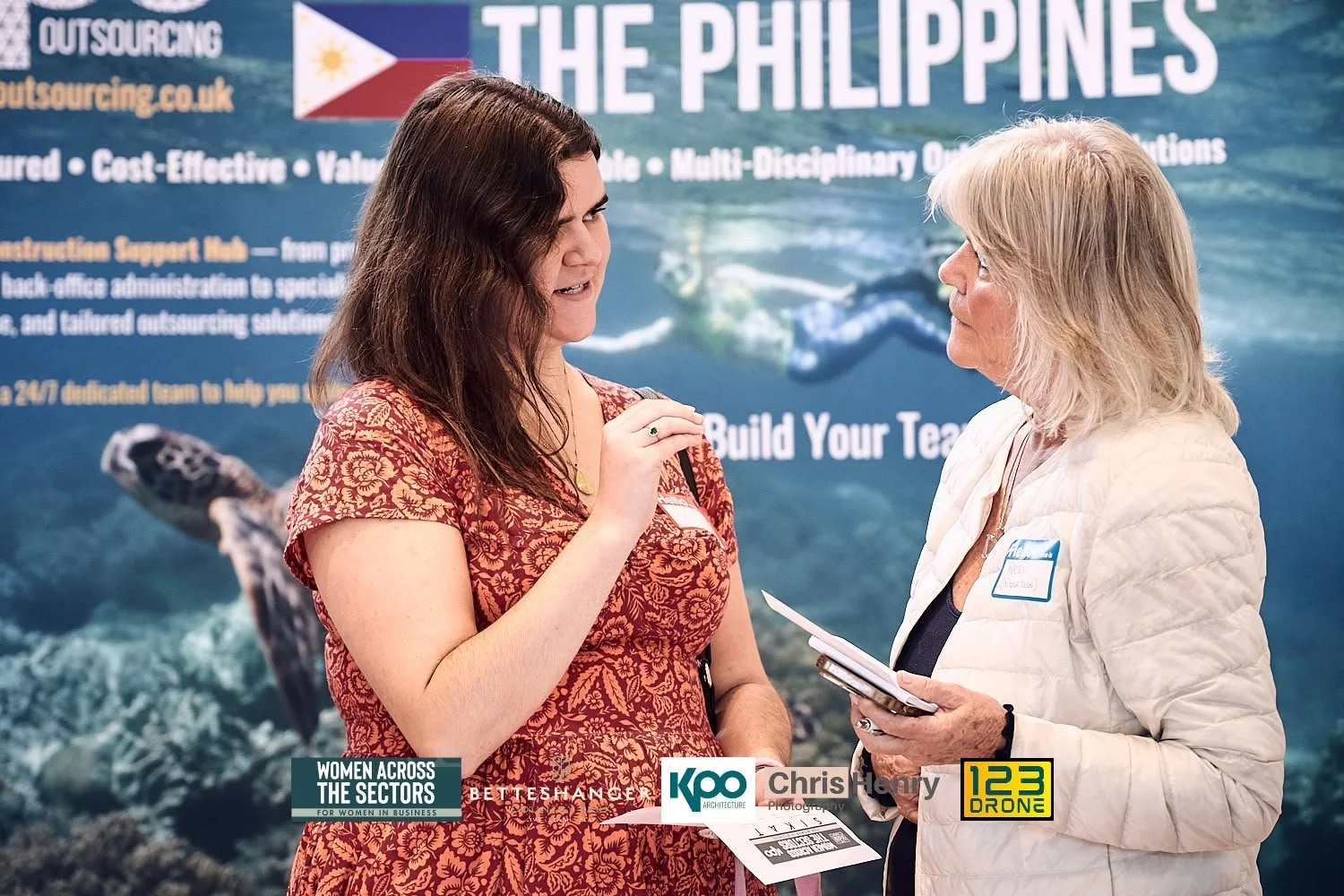 Two women engaged in conversation at a conference or event booth with a banner reading 'The Philippines' in the background. The woman on the left, with dark hair and a patterned dress, gestures as she talks. The woman on the right, with blonde hair a